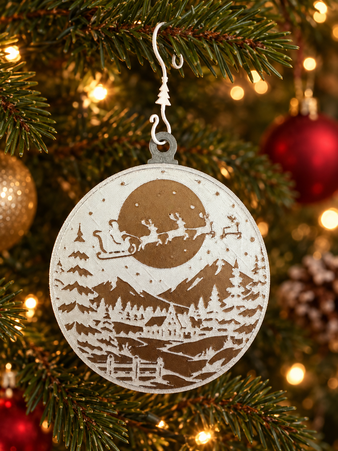 Christmas tree ornament with a cutout scene of Santa riding his sleigh with reindeer, flying over mountains, trees, a house, and a pond, hanging on a green Christmas tree decorated with lights.