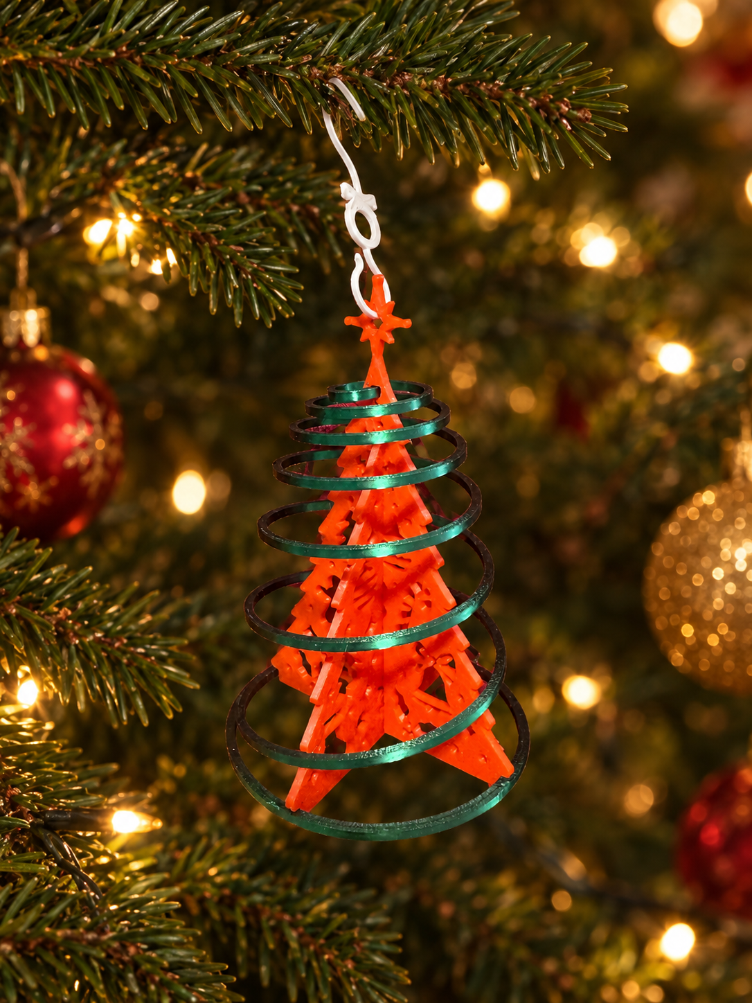 Decorative Christmas tree ornament resembling the Eiffel Tower, hanging on a Christmas tree with blurred lights and red ornaments in the background.