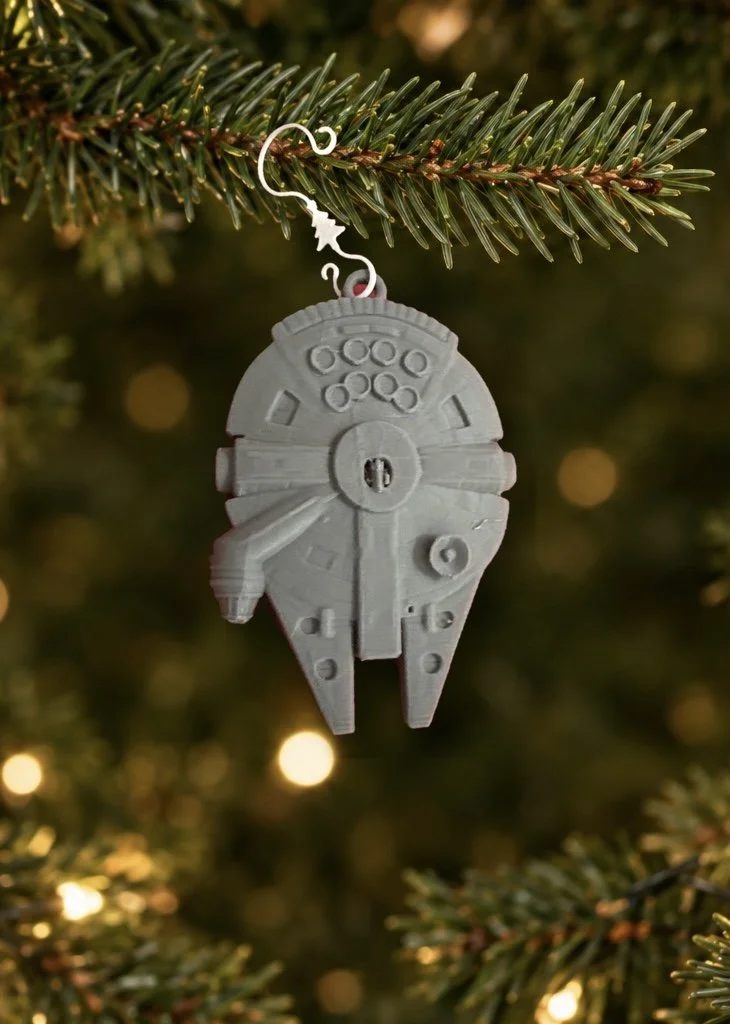 A Star Wars Millennium Falcon ornament hanging from a Christmas tree branch with blurred lights in the background.