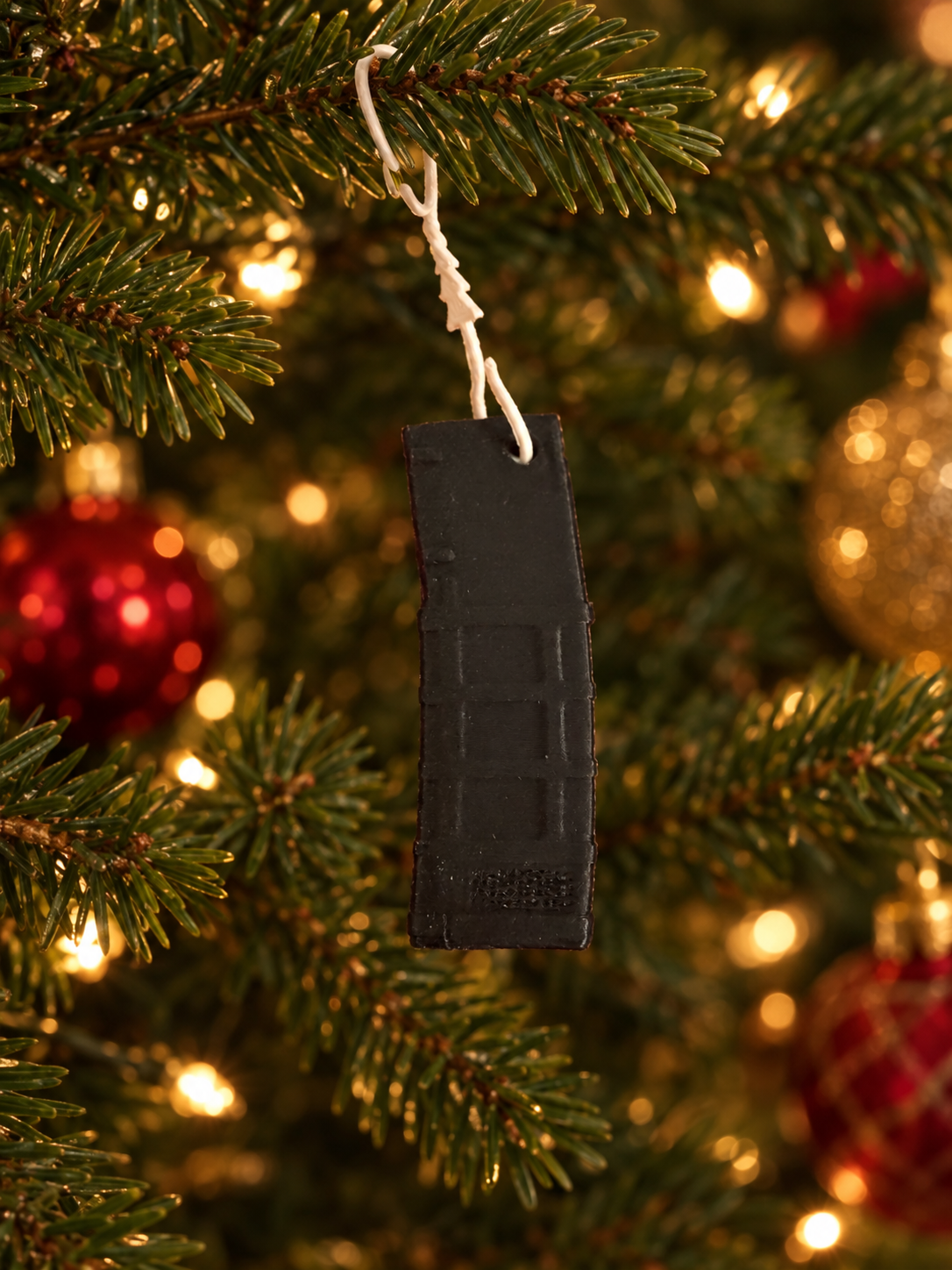 A close-up of a Christmas tree ornament shaped like a black cigarette lighter, hanging from a branch of a decorated Christmas tree with blurred lights and ornaments in the background.