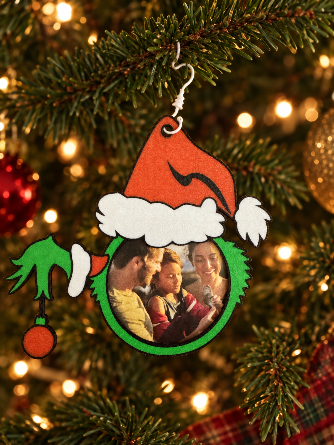 A Christmas tree ornament with a photo of a family, decorated with a red Santa hat, green leaves, and a red ornament, hanging on a Christmas tree with lights.