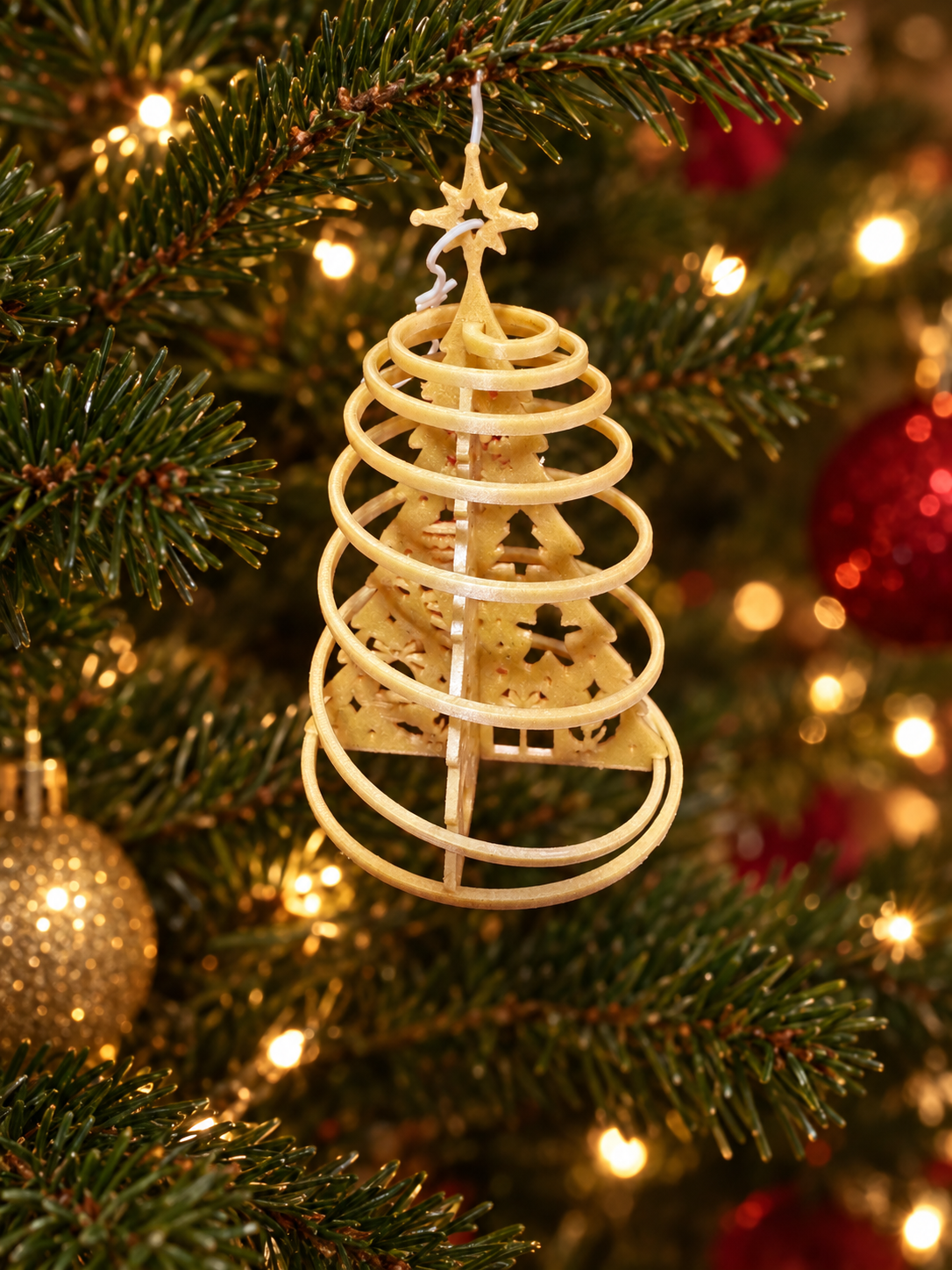 A Christmas tree ornament shaped like a spiral, with a star on top, hanging from a Christmas tree decorated with lights and red ornaments.