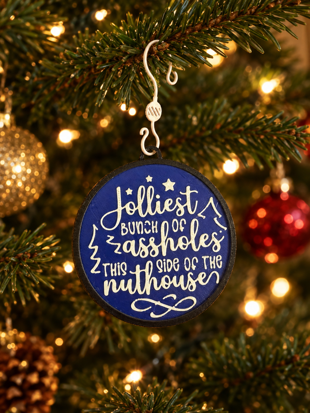 A Christmas tree ornament with humorous text, hanging on a decorated tree with lights and other ornaments in the background.
