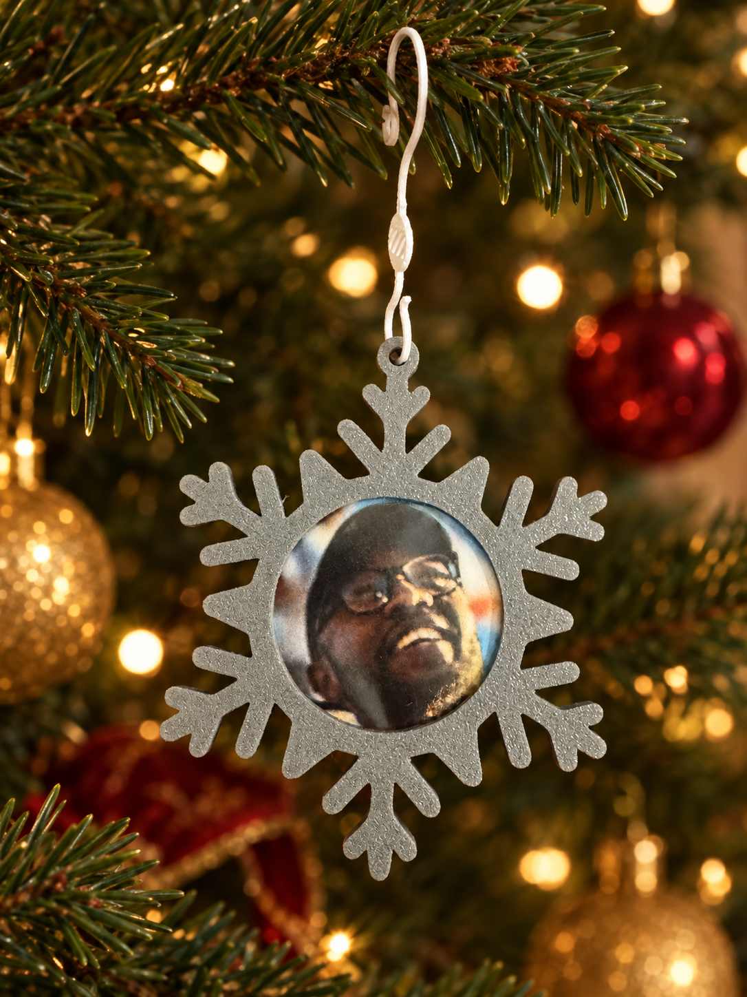 Decorative snowflake-shaped ornament hanging on a Christmas tree featuring a photograph of a man with glasses and a wide smile, surrounded by other blurred Christmas ornaments and warm lights.