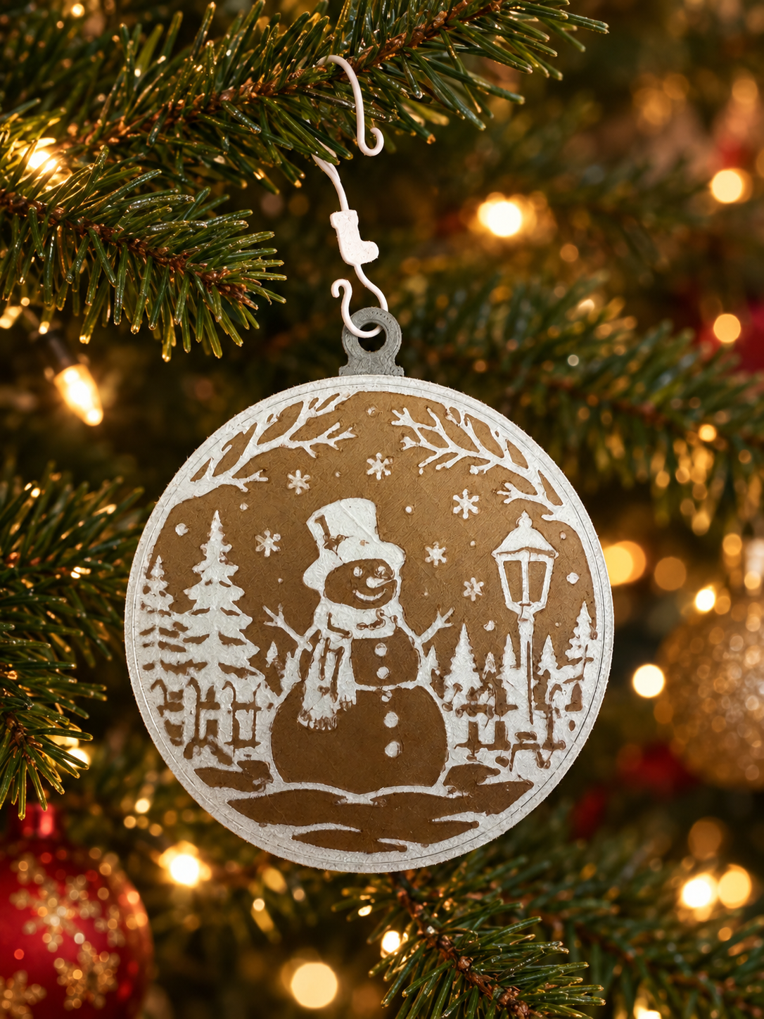 Christmas tree ornament depicting a snowman wearing a hat and scarf, surrounded by trees, a lamppost, and snowflakes.