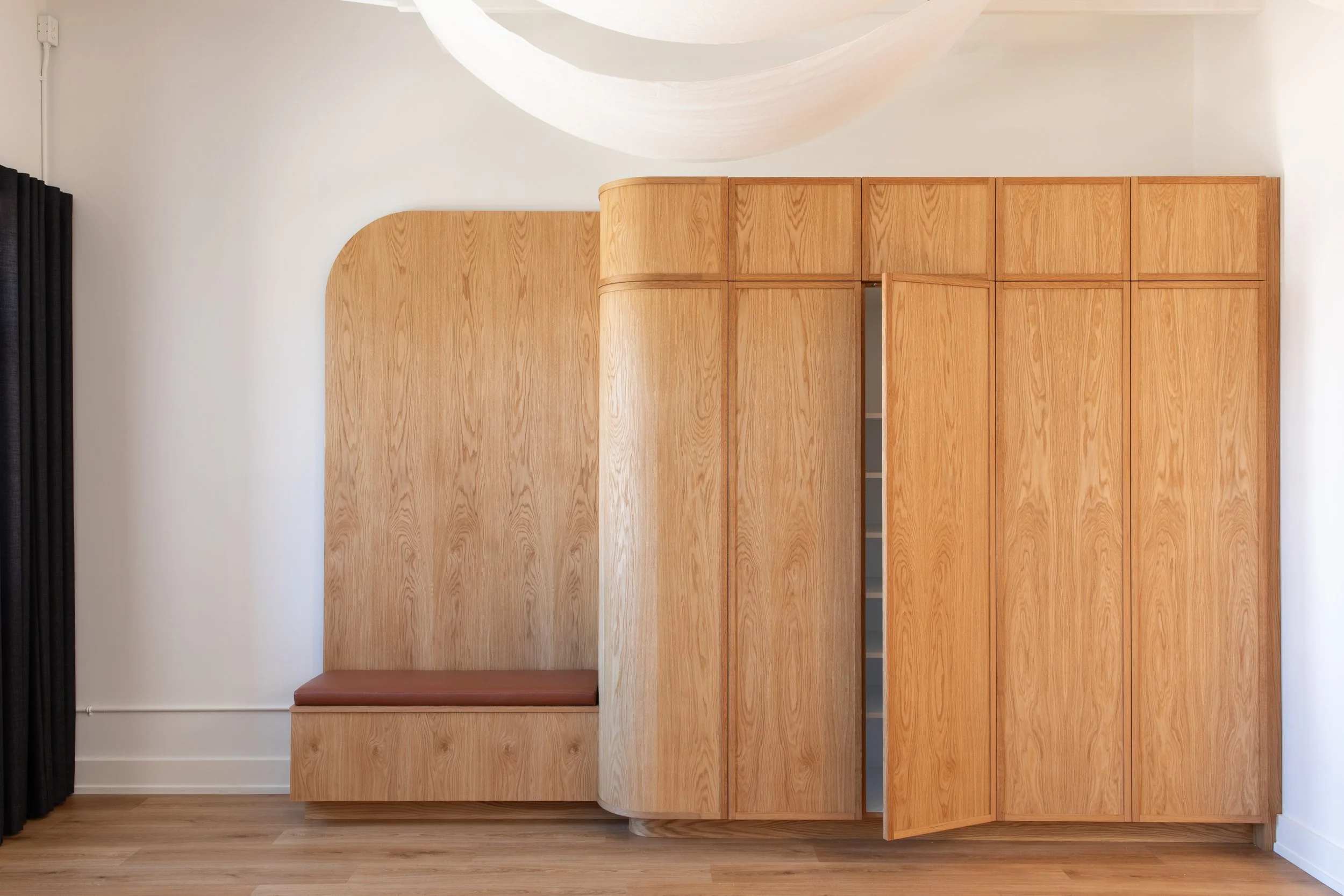 Full view of custom oak storage cabinetry at Wonderlit Studios, with an integrated upholstered bench, rounded cabinet edges and an open door revealing interior shelving.   Copy all image SEO