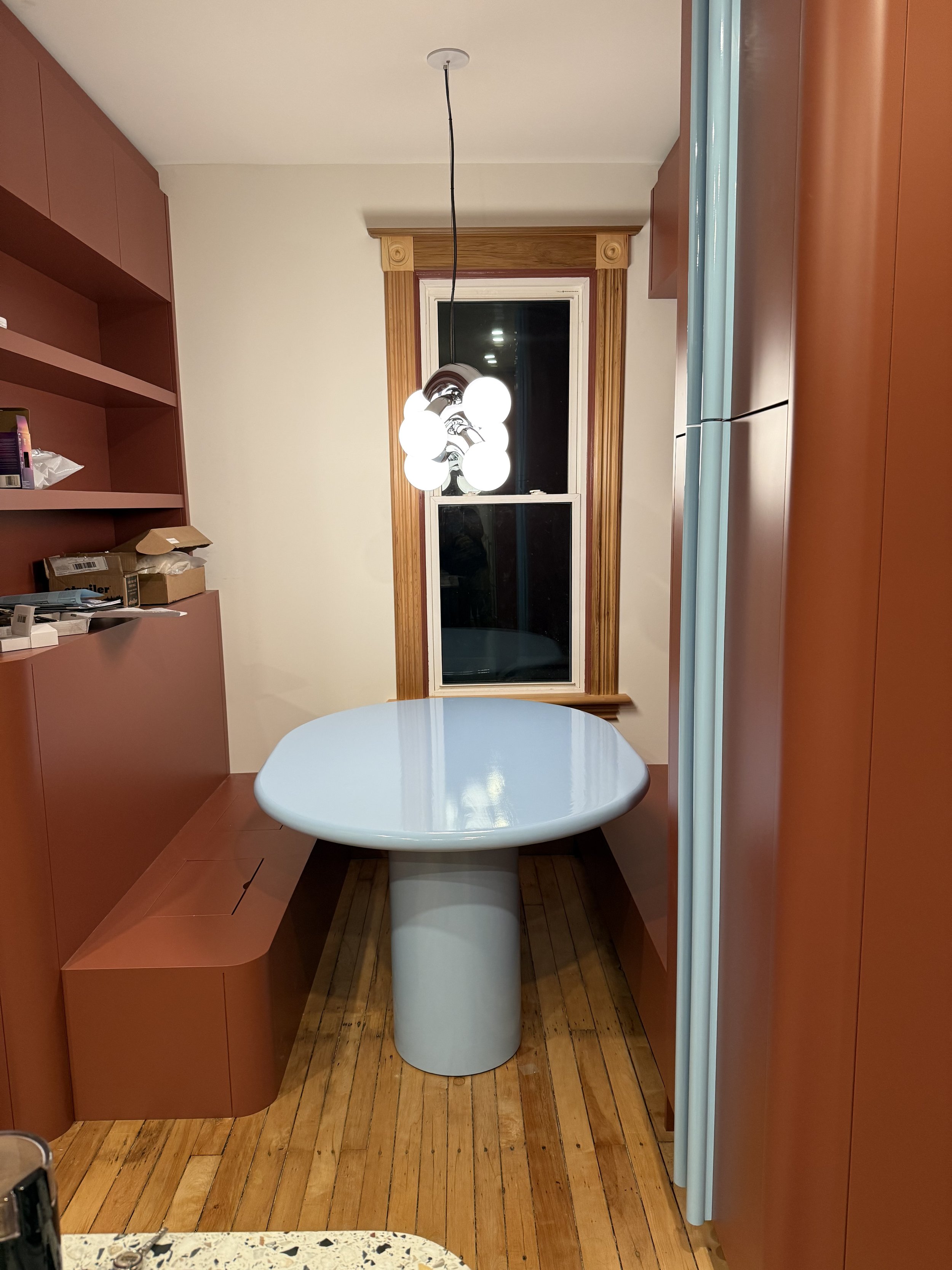 Front view of the custom pale blue breakfast table installed between terracotta cabinetry and built-in banquette seating, with original wood flooring and a globe pendant light.