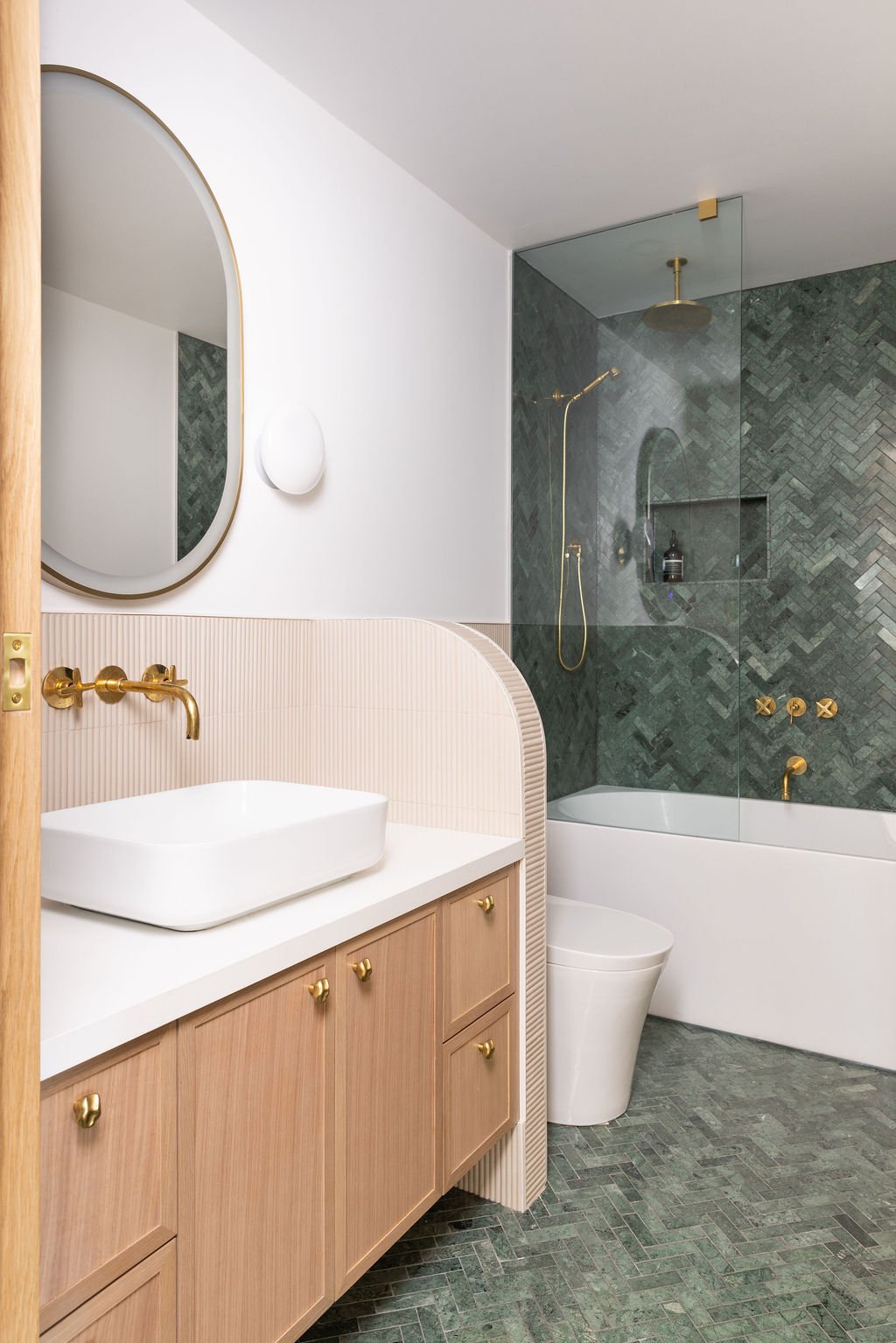 Residential bathroom featuring a custom oak vanity, arched mirror, white vessel sink and glass shower enclosure with green stone tile.