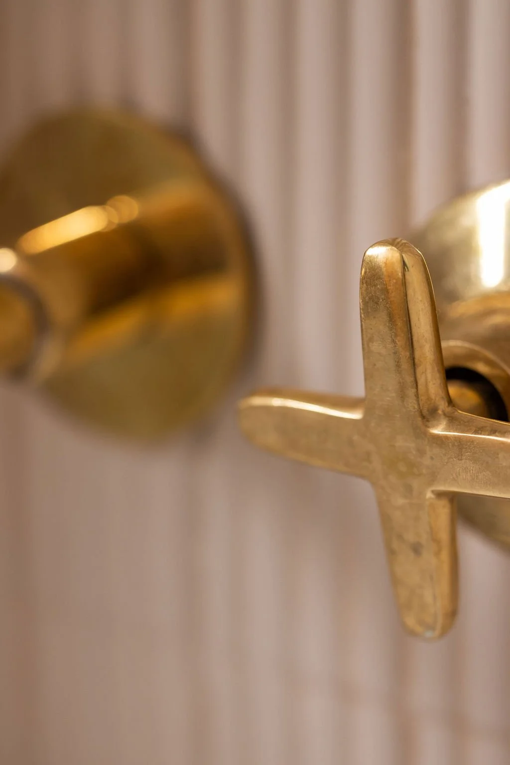 Close-up of aged brass cross-handle bathroom fixtures against pale vertically fluted tile.