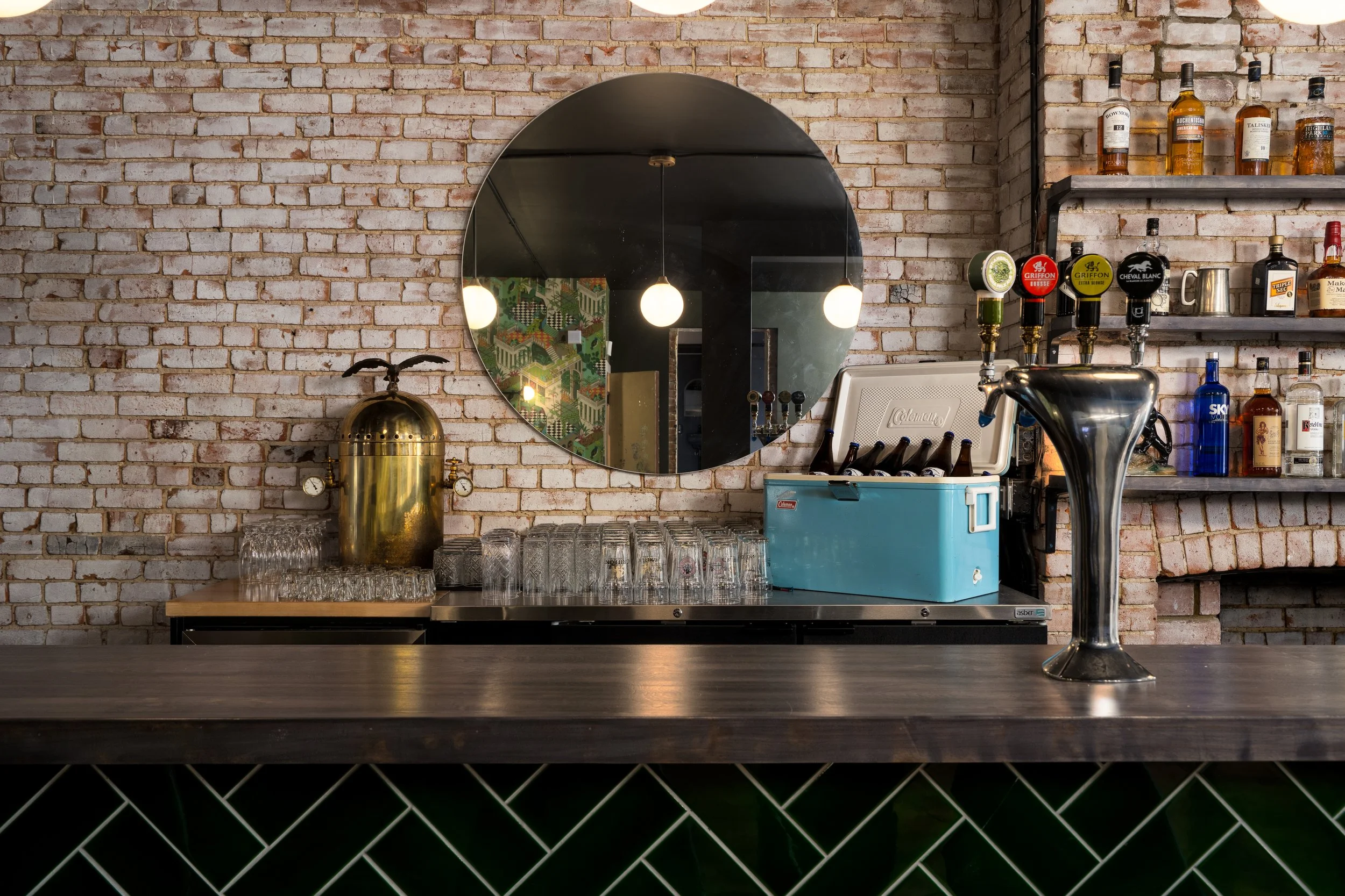 Bar interior at Turbo Haüs in Montréal featuring exposed brick, a circular mirror, dark wood countertop and deep green herringbone tile.