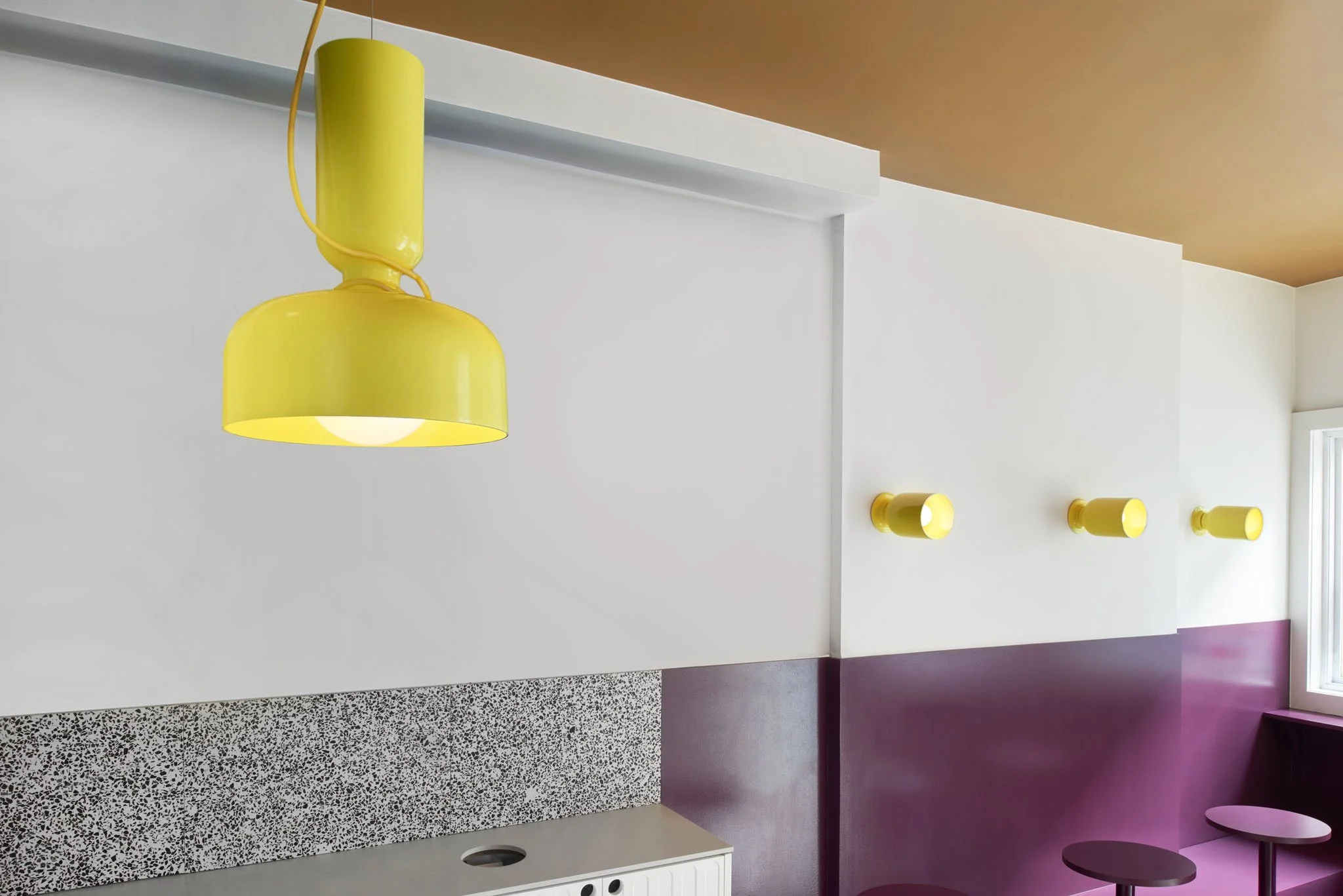 Close-up of a yellow pendant and matching wall lights against white architectural panels, plum walls and a speckled backsplash.