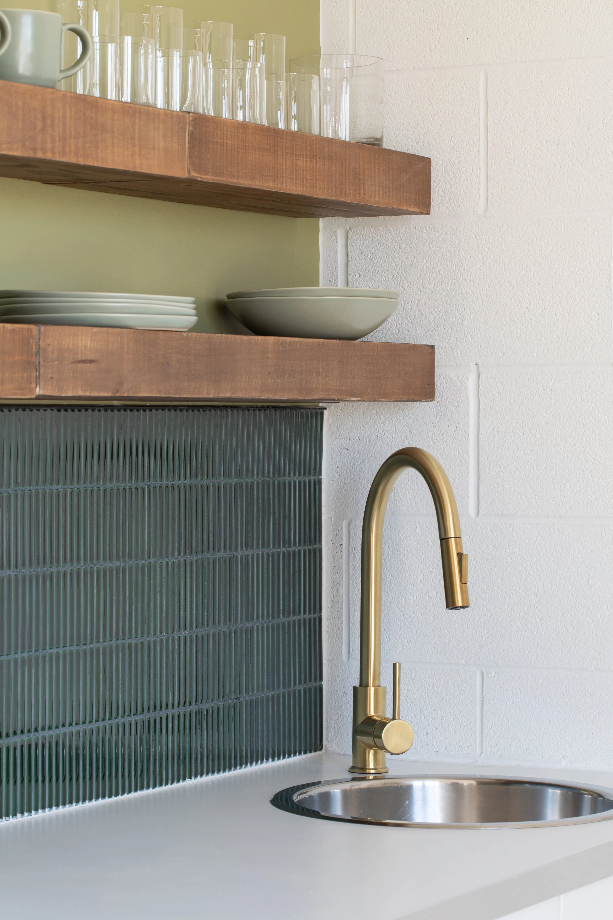 Studio kitchenette with dark green vertical tile, floating wood shelves, white countertop and brushed brass faucet.