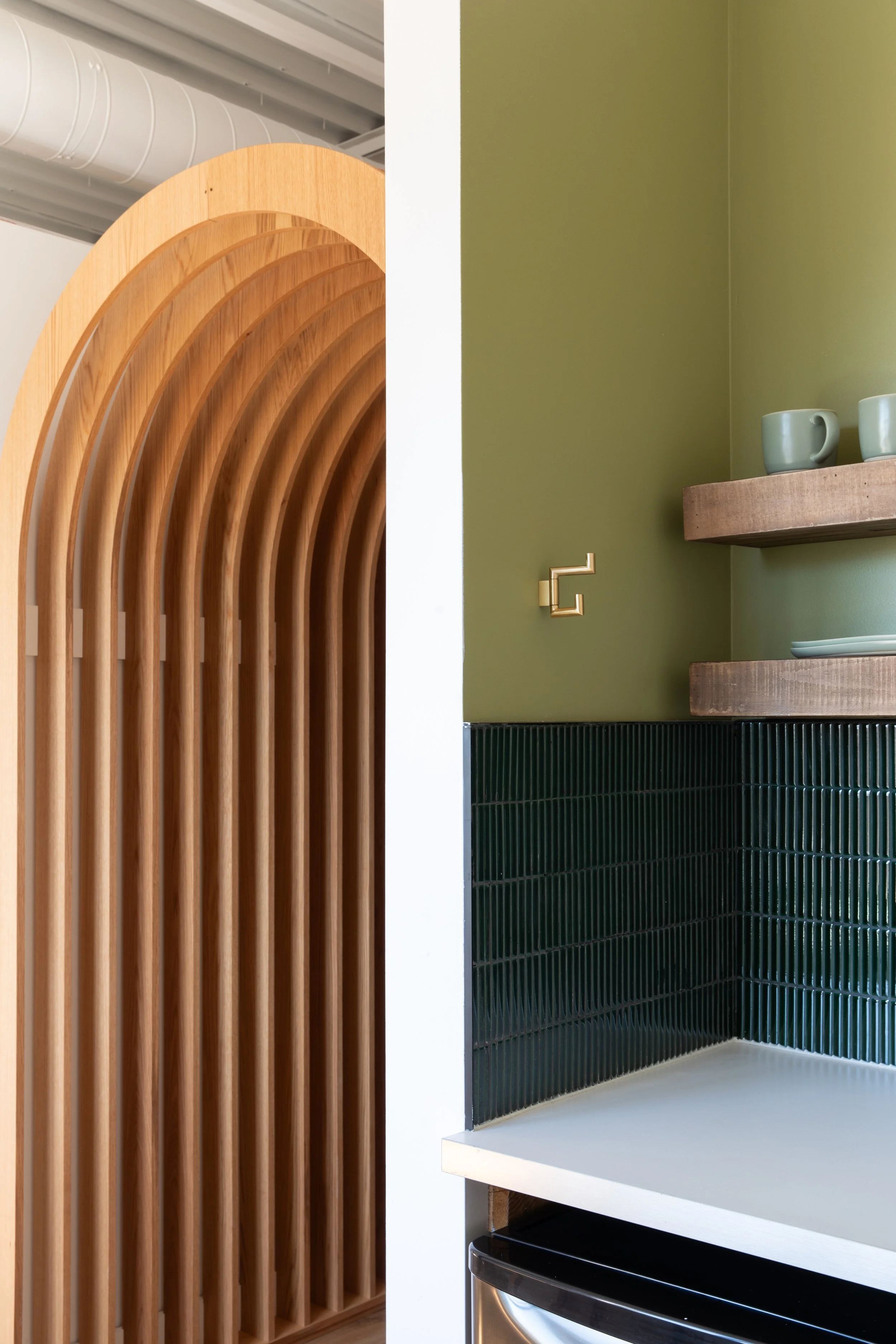 Compact kitchenette with olive green walls, dark green tile backsplash, floating wood shelves and white countertop.