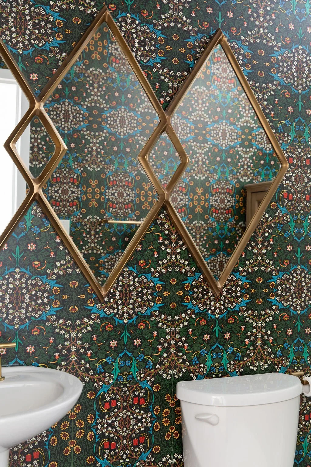Geometric wood-framed mirrors against richly patterned floral wallpaper above a white bathroom sink.