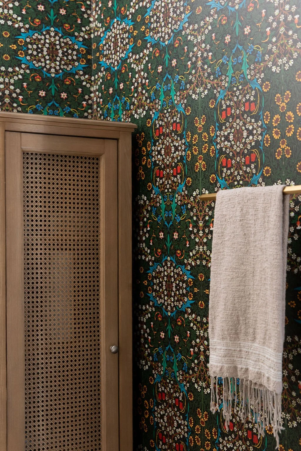 Colourful floral wallpaper, a wood cabinet with woven cane panel and a hanging towel in the Wonderlit Studios bathroom.