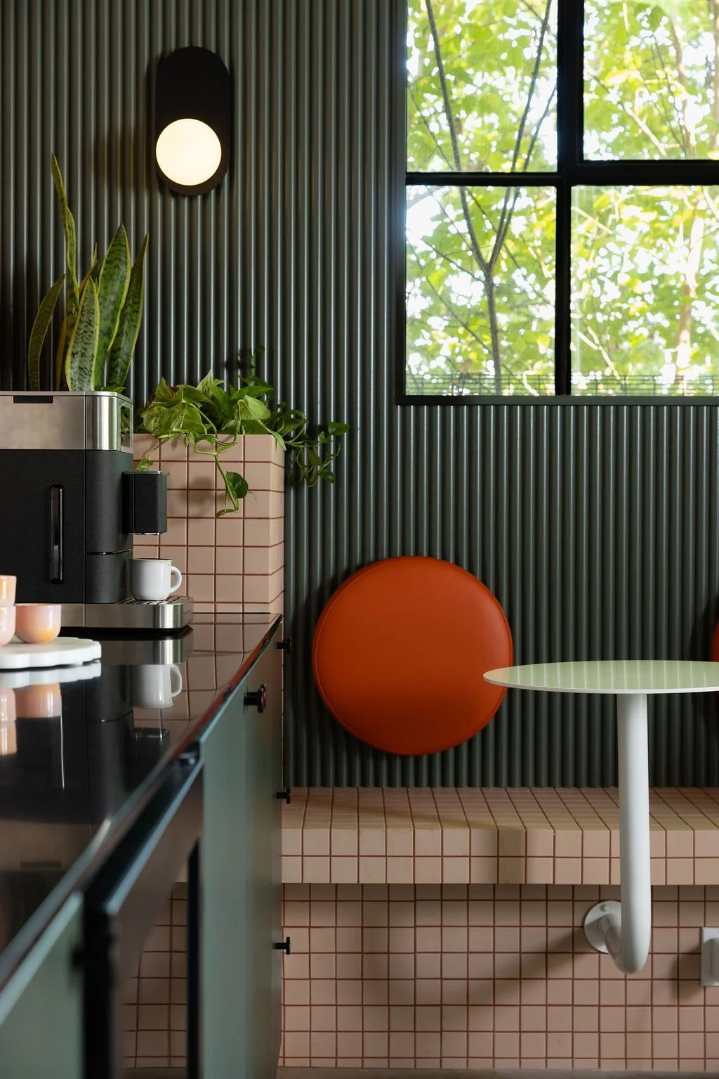 Orange circular upholstered backrest against dark green fluted wall panels, with a pink tiled bench and white café table.