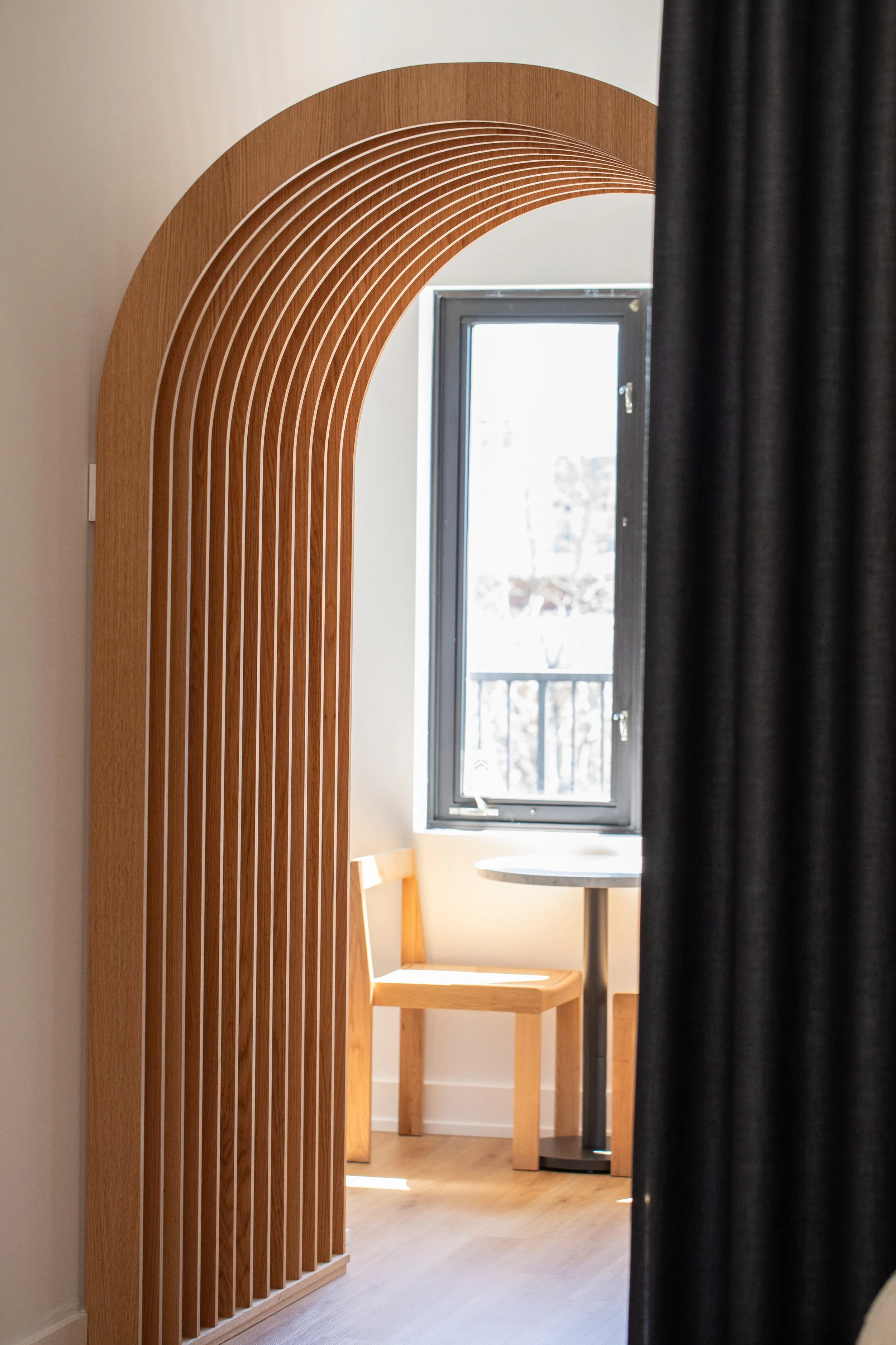Curved wood archway framing a sunlit seating nook with a small table and chair beside a window.