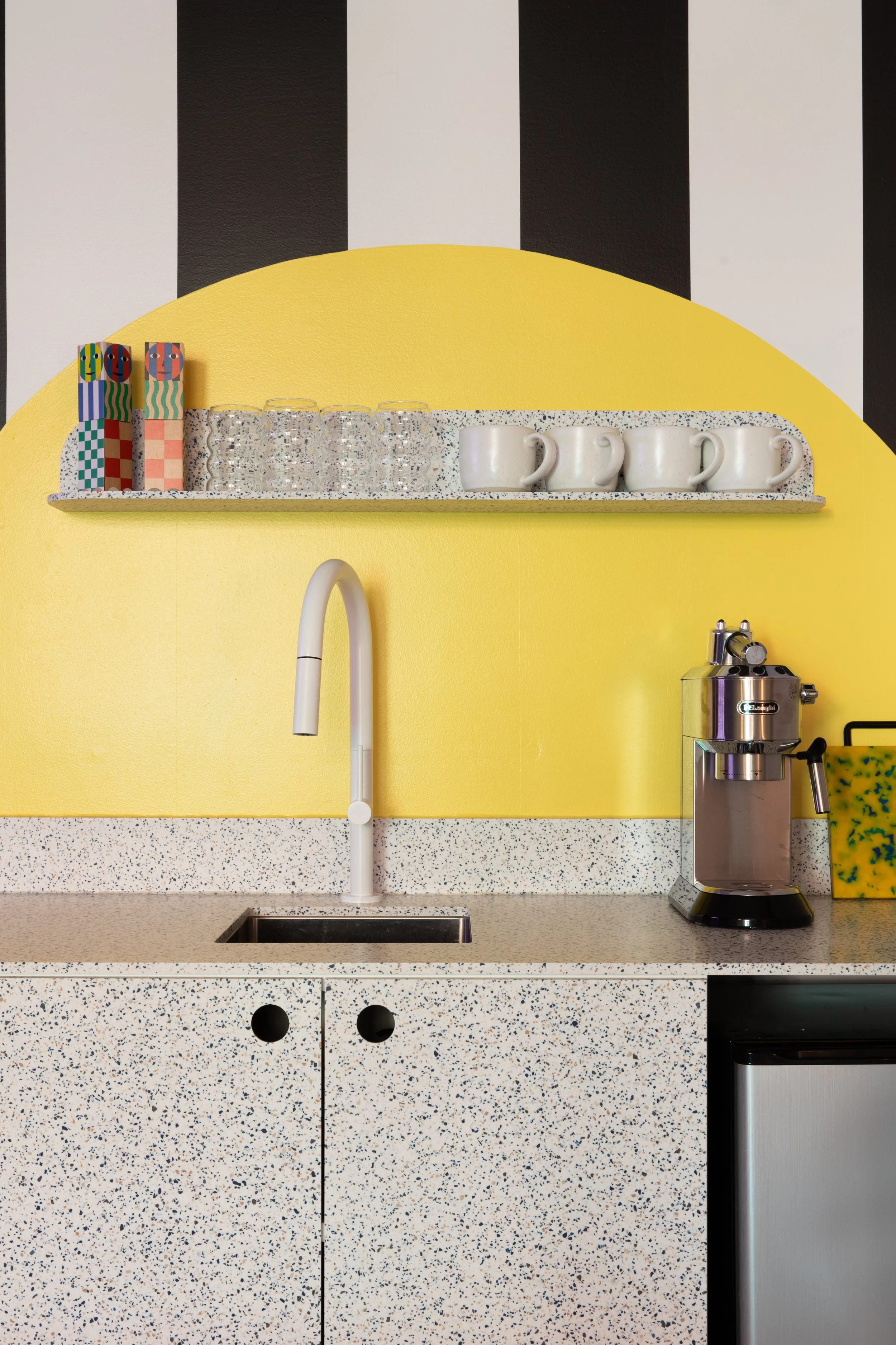 Yellow arched kitchenette with a speckled countertop, floating shelf, white mugs and stainless steel coffee equipment.