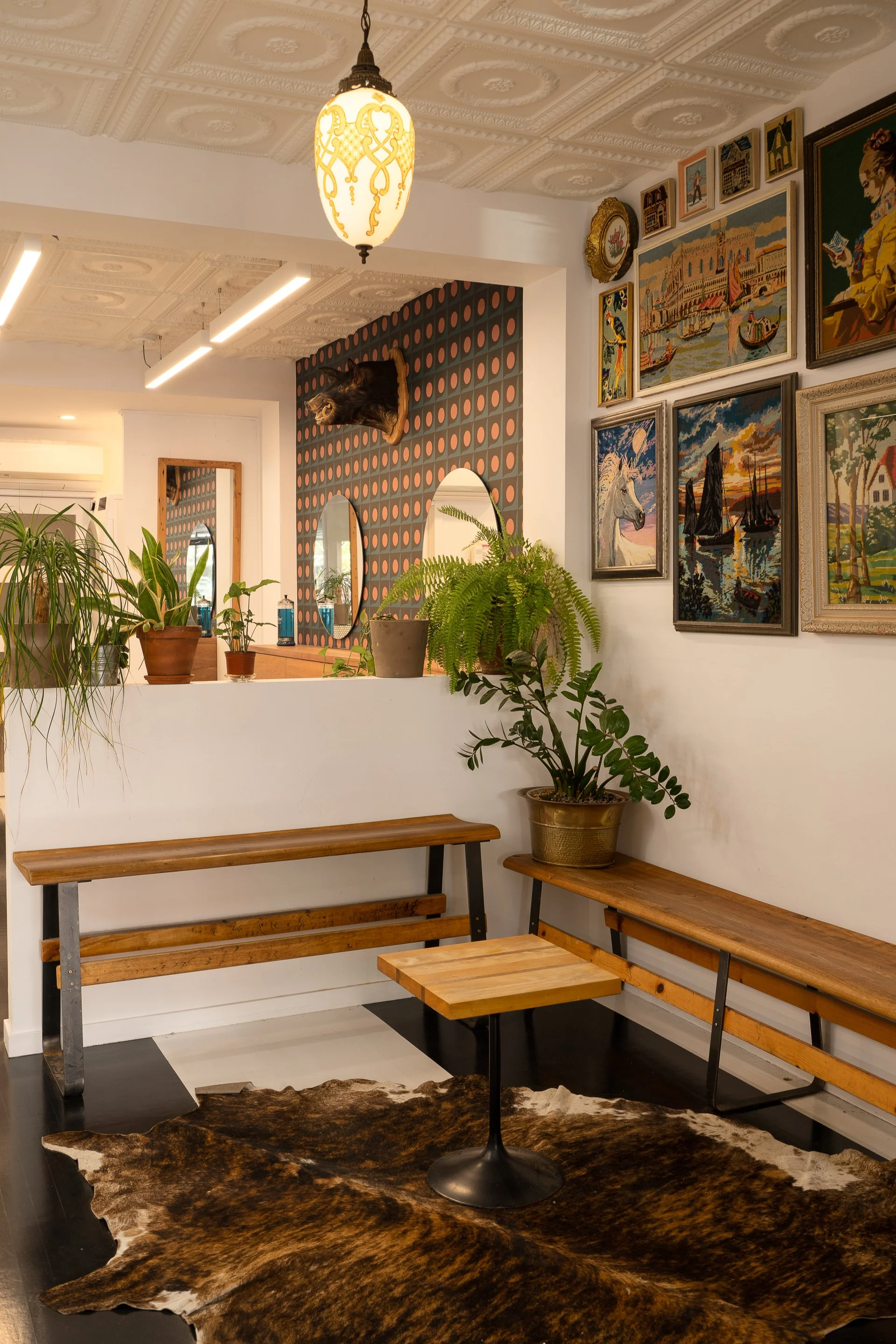 Two Horses salon waiting area featuring wooden benches, vintage framed artwork, indoor plants and black-and-white striped flooring.