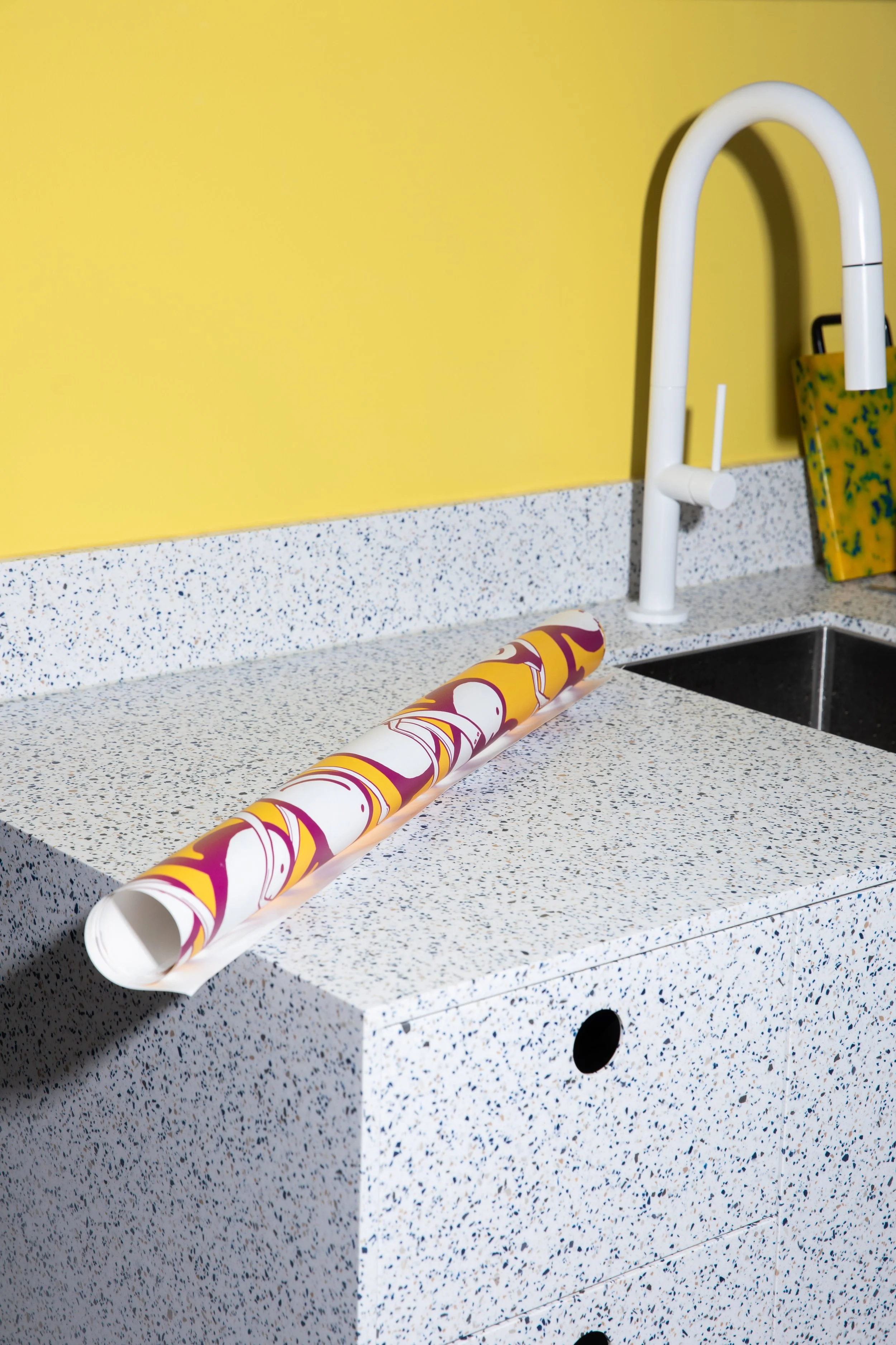 Pink, yellow and white patterned wallpaper roll displayed on a speckled countertop beside a white faucet.