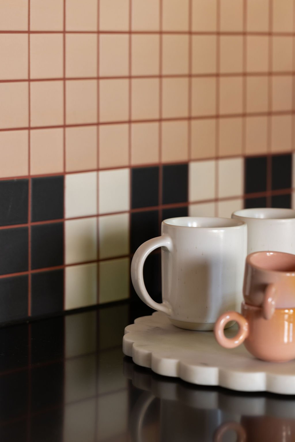 Pale pink and charcoal grid tiles behind a black countertop, with ceramic mugs arranged on a sculptural white tray.