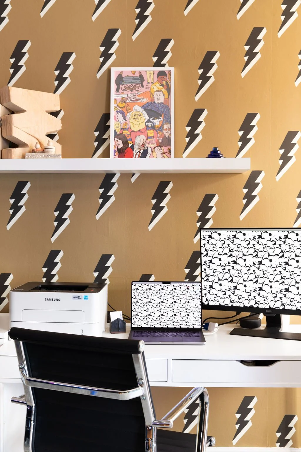 Creative studio workstation with white desk, black office chair, floating bookshelf and graphic lightning bolt wallpaper.