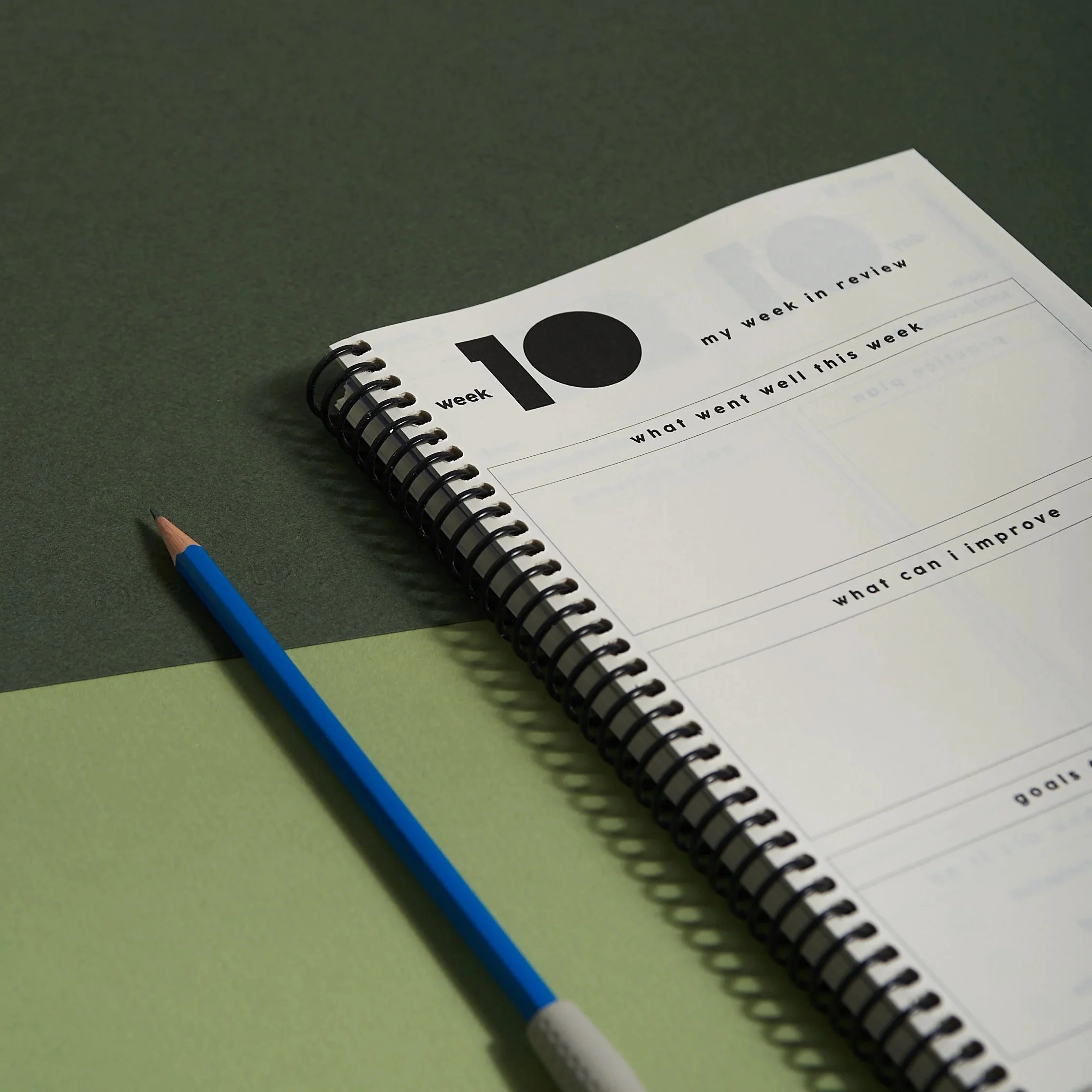 Open spiral-bound Notabl. practice journal on a green surface with a blue pencil, showing a weekly review page for reflection and goal setting.