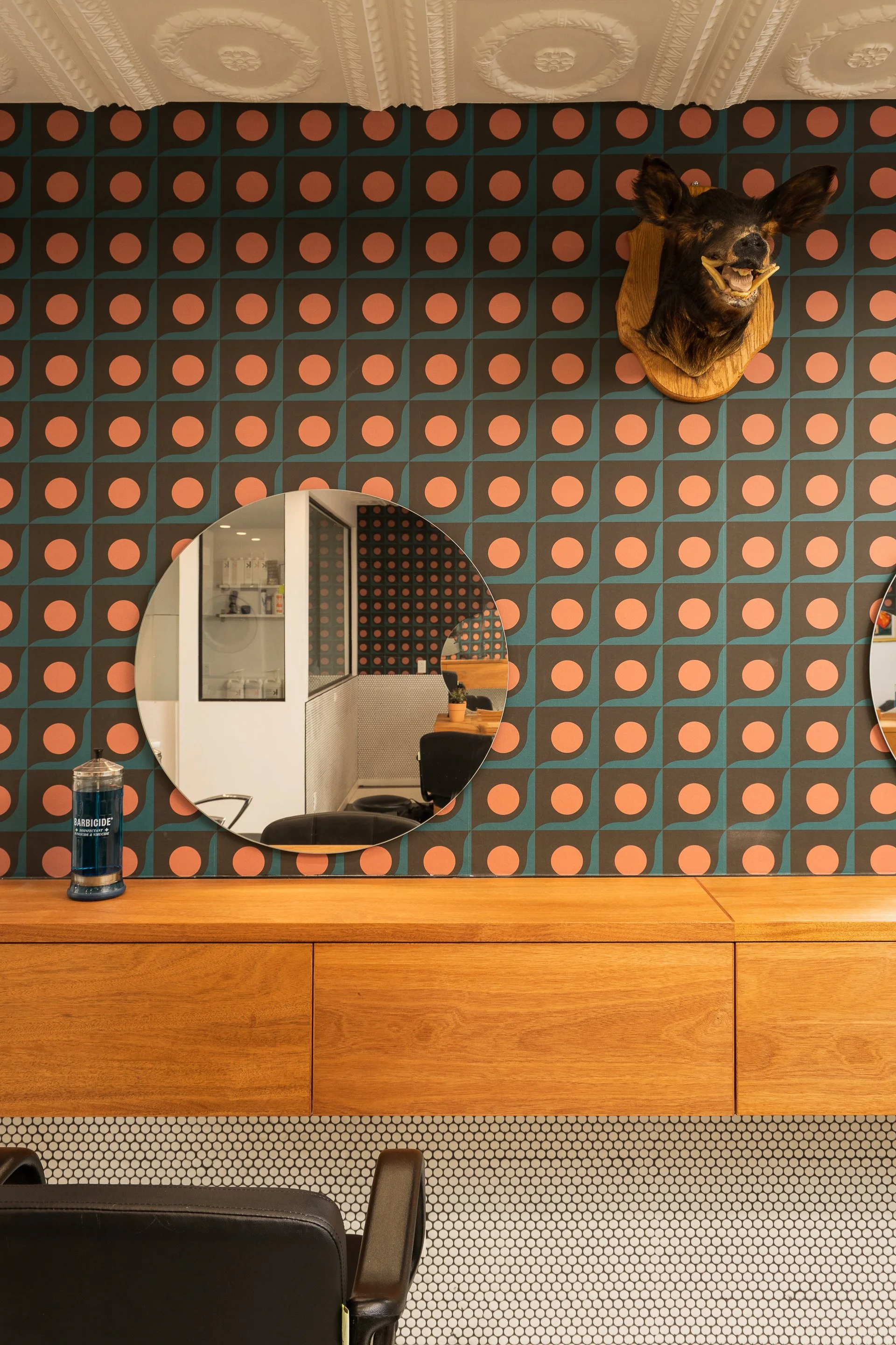 Custom wood salon styling station with a circular mirror, orange-and-black patterned wallpaper and vintage wall-mounted decor at Two Horses.