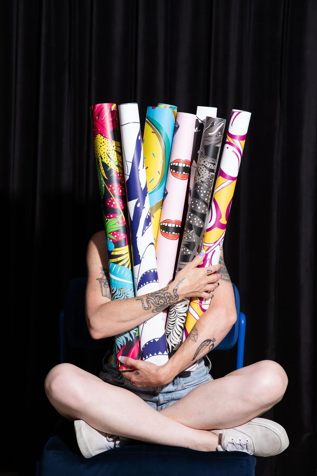 Editorial photograph of a seated designer holding colourful rolls of original flodström wallpaper against a black curtain.