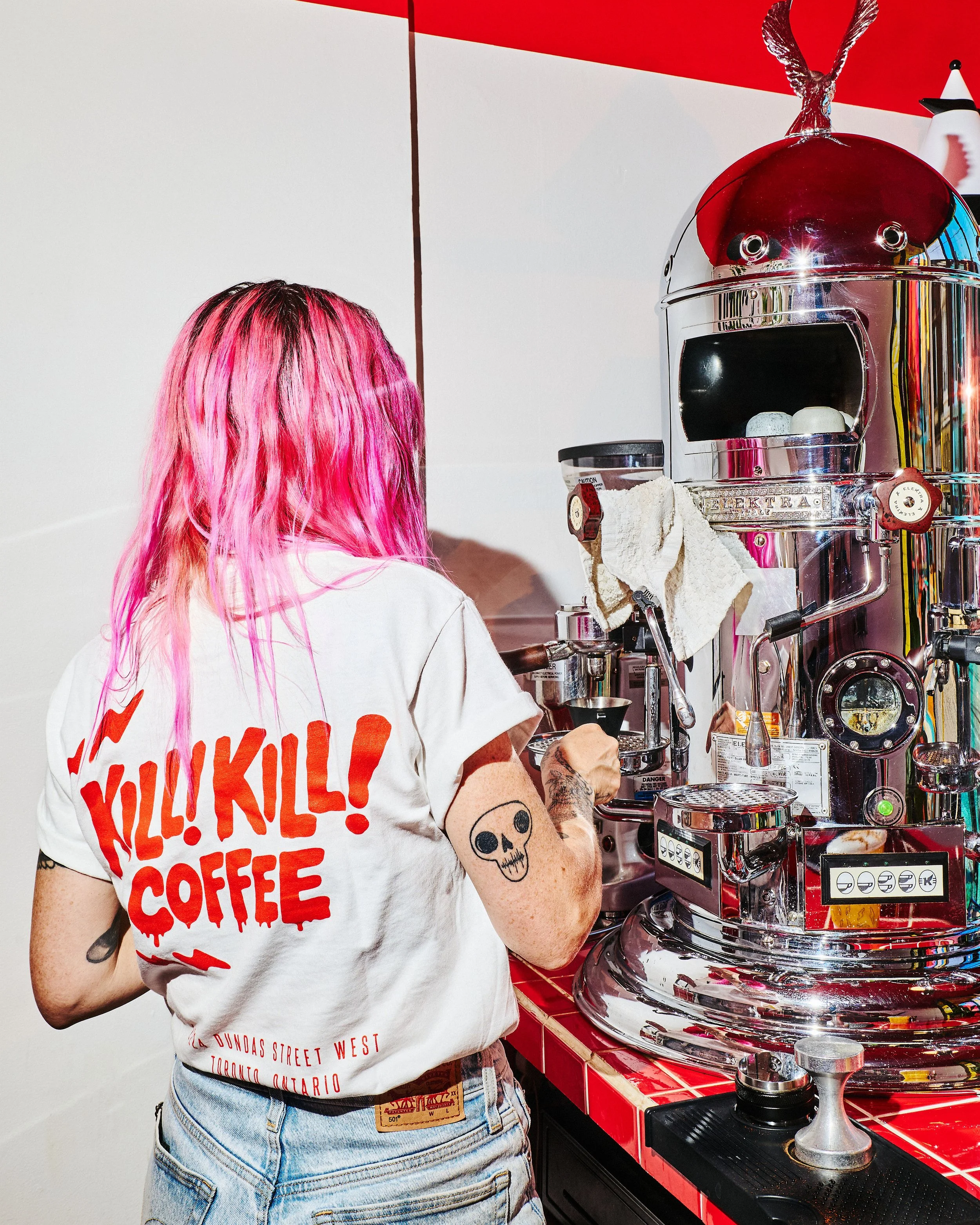 Barista wearing a white Kill! Kill! Coffee T-shirt with a red logo while working at a chrome espresso machine.    Copy all image names and alt text