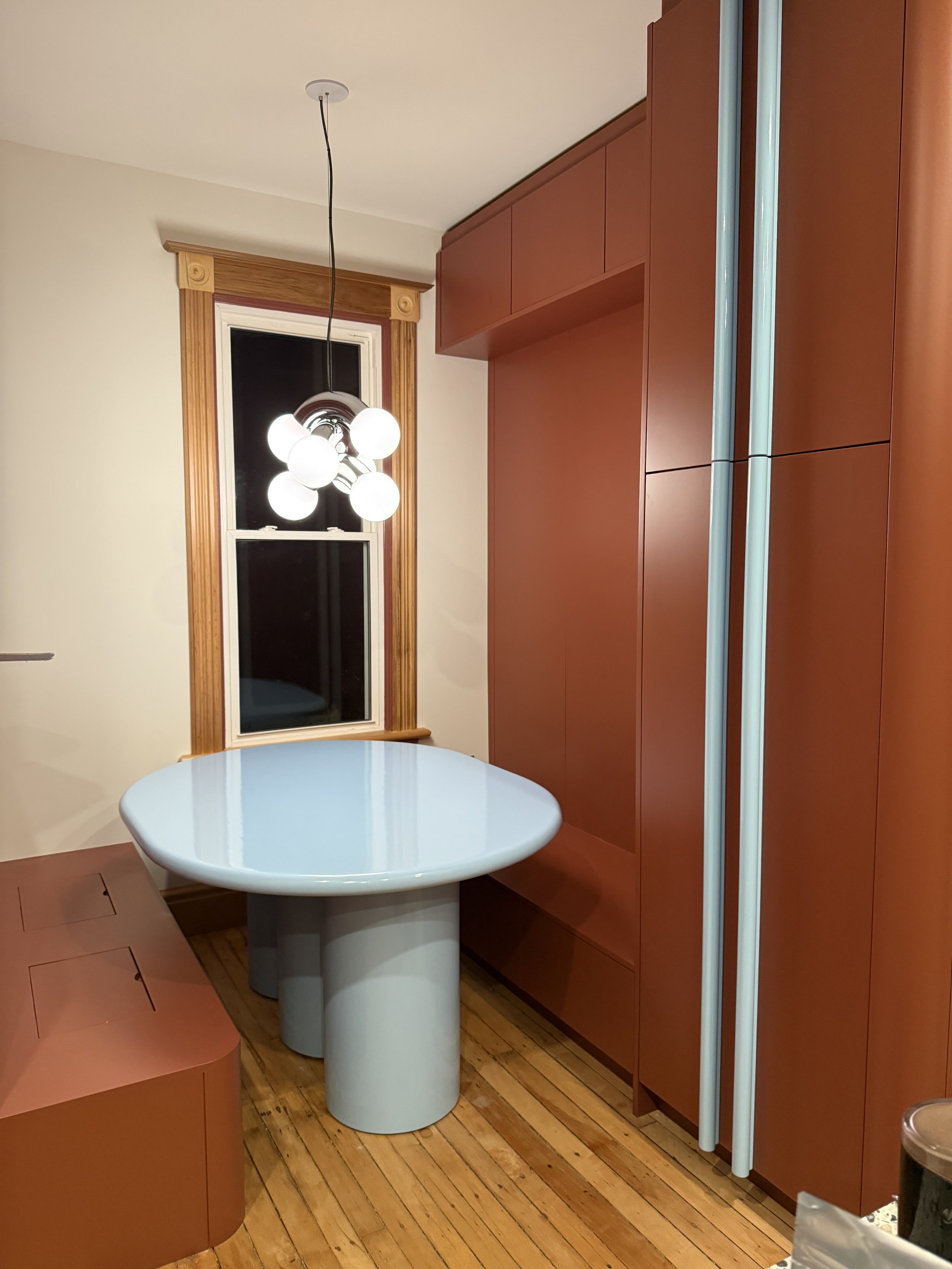 Custom pale blue breakfast table with a rounded sculptural top and three cylindrical legs, installed beside a terracotta built-in banquette in a residential kitchen.