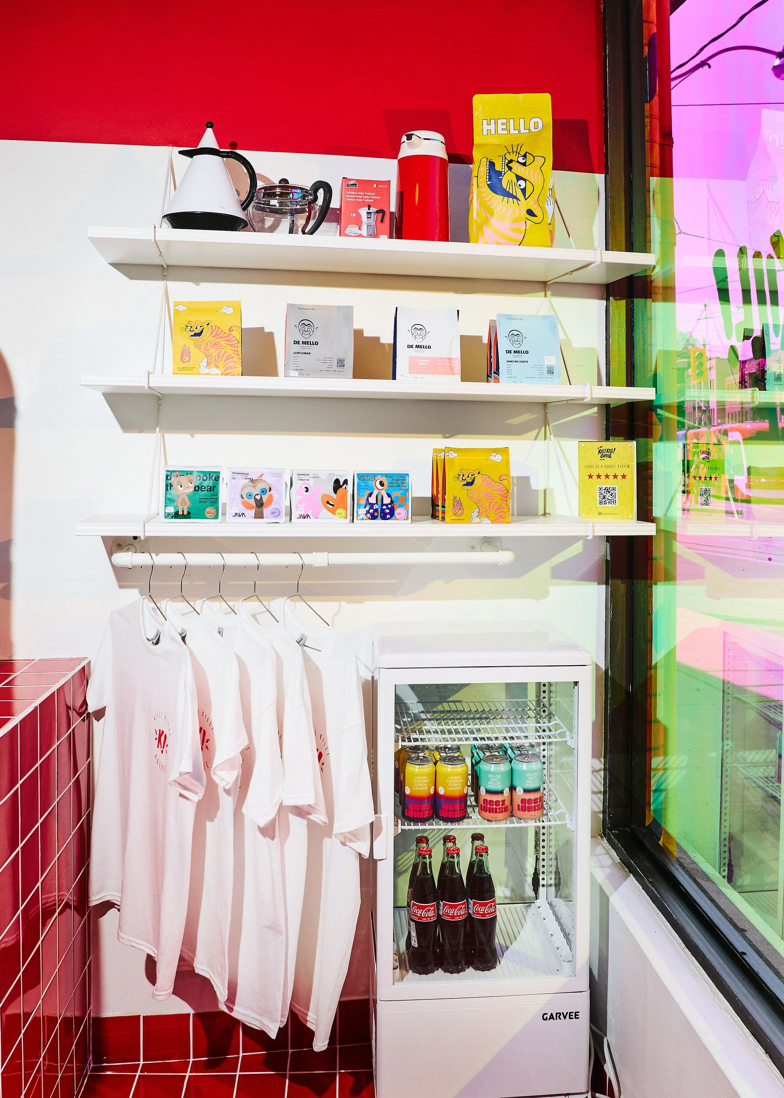 Café retail display with colourful coffee bags, branded T-shirts and a drinks fridge beside the front window.
