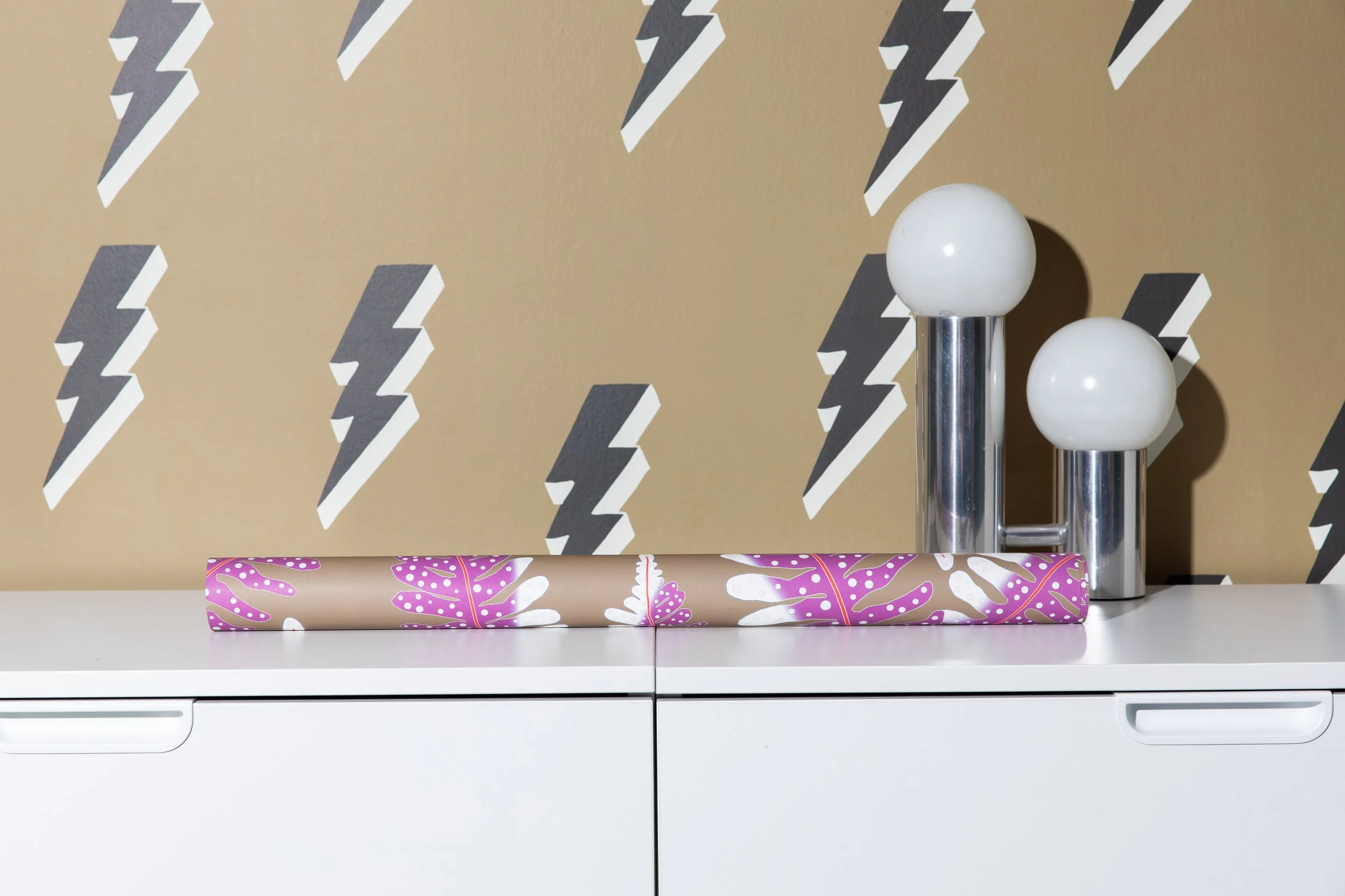 Tan wallpaper with repeating black-and-white lightning bolts behind white storage cabinetry and sculptural chrome table lamps.