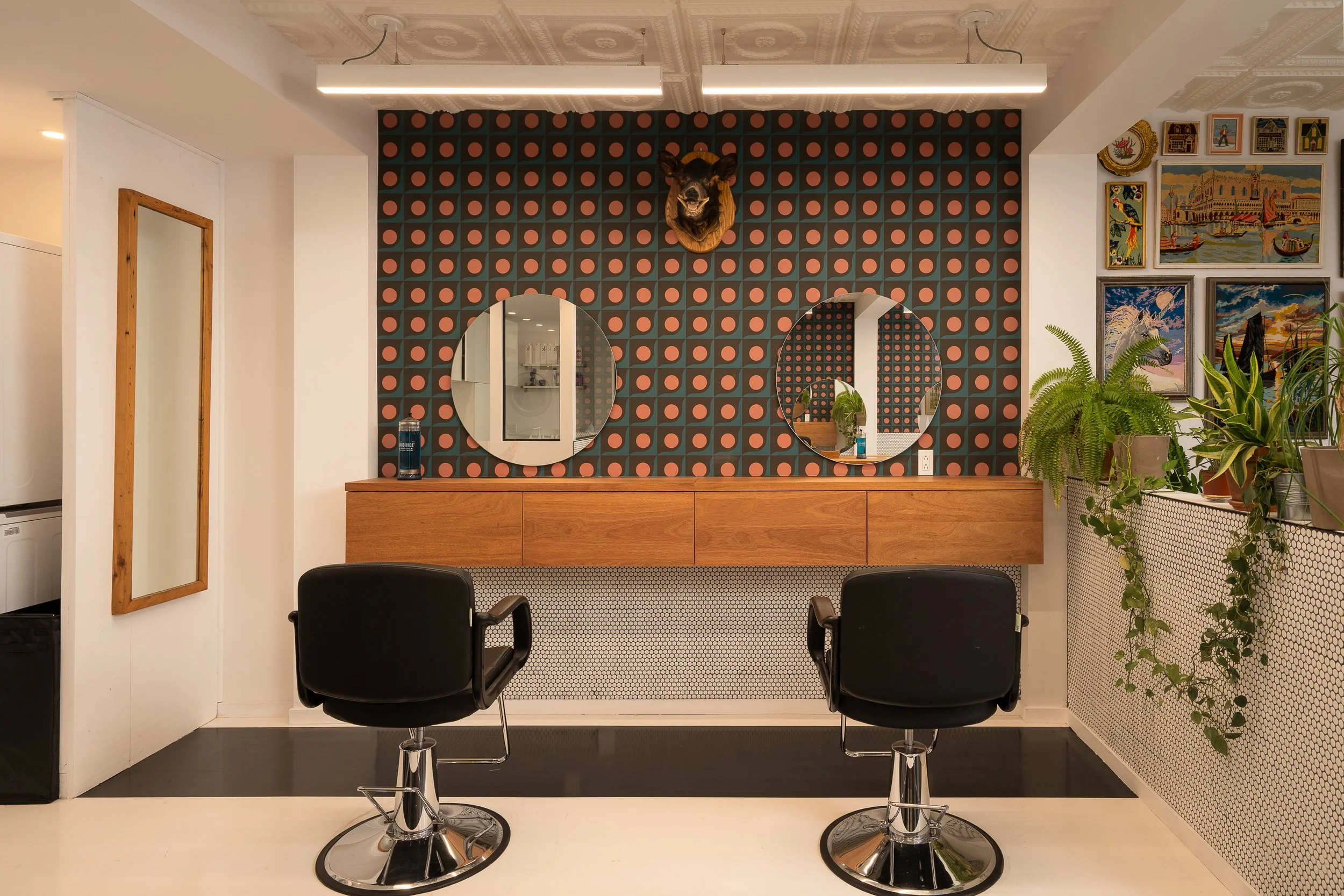 Two Horses hair salon styling stations featuring custom wood cabinetry, circular mirrors, patterned wallpaper and black salon chairs.
