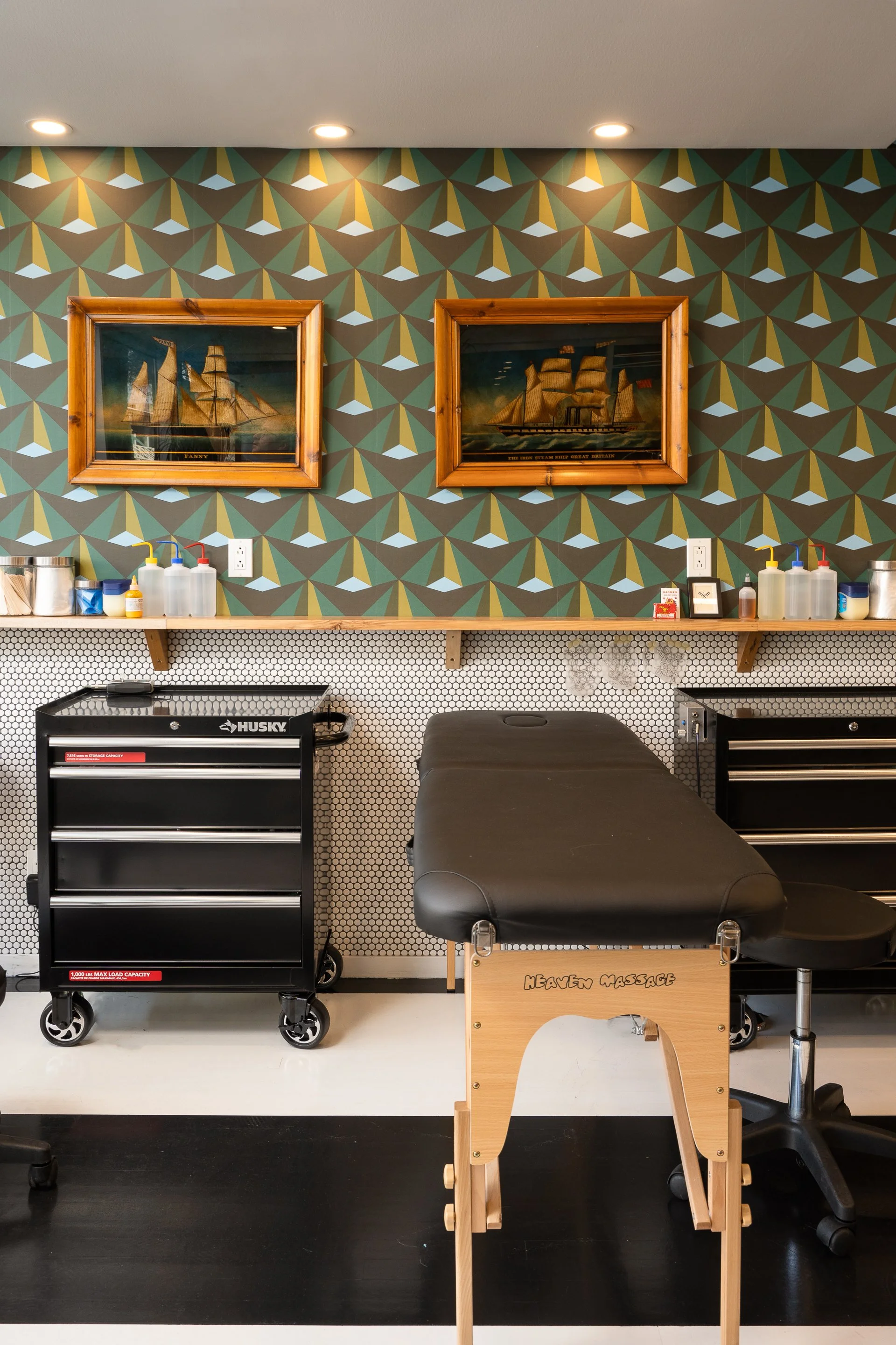 Tattoo workstation featuring a black treatment bed, geometric wallpaper, framed artwork and black storage cabinets.