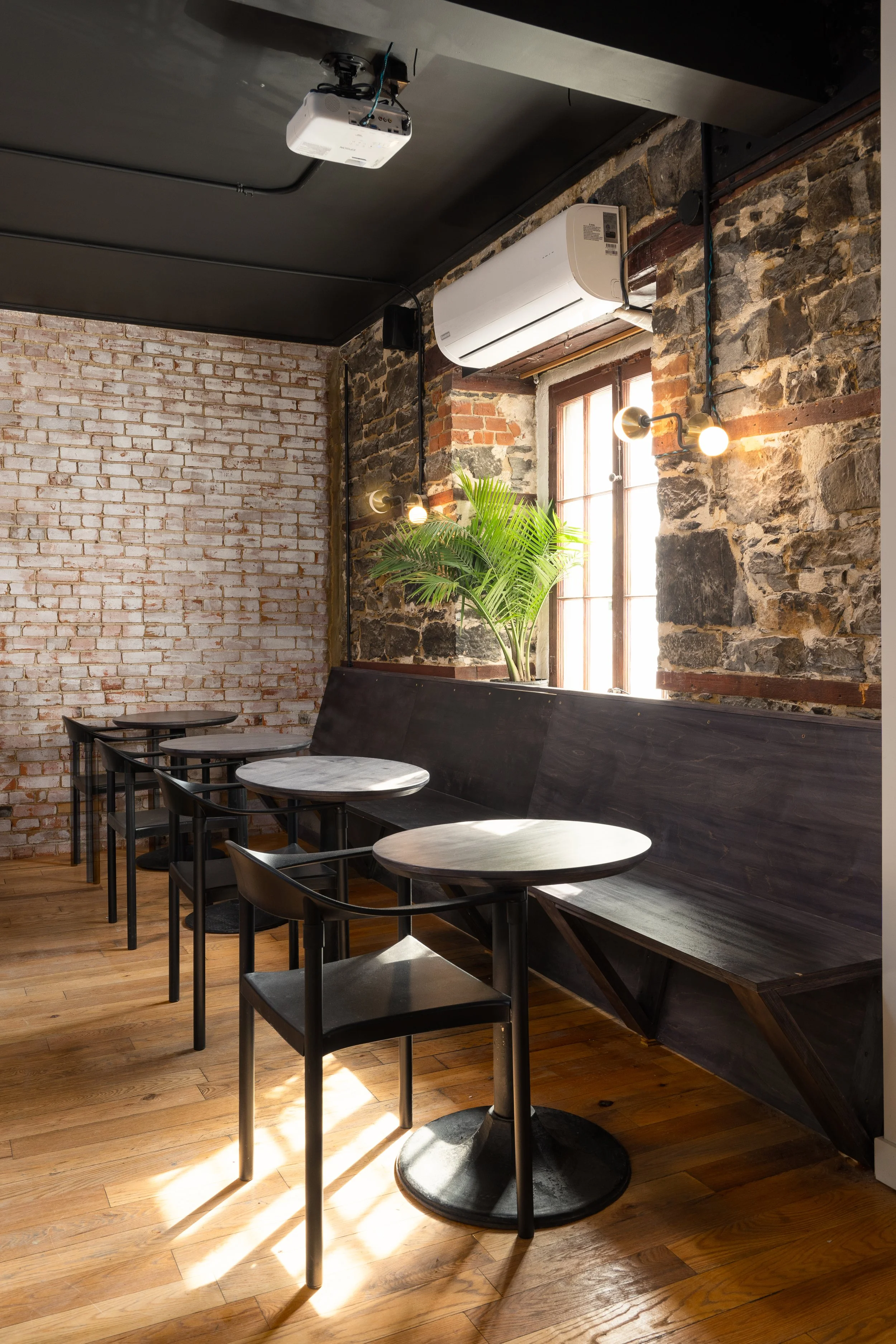 Sunlit seating area with dark timber banquettes, black café tables, exposed masonry and wall sconces.