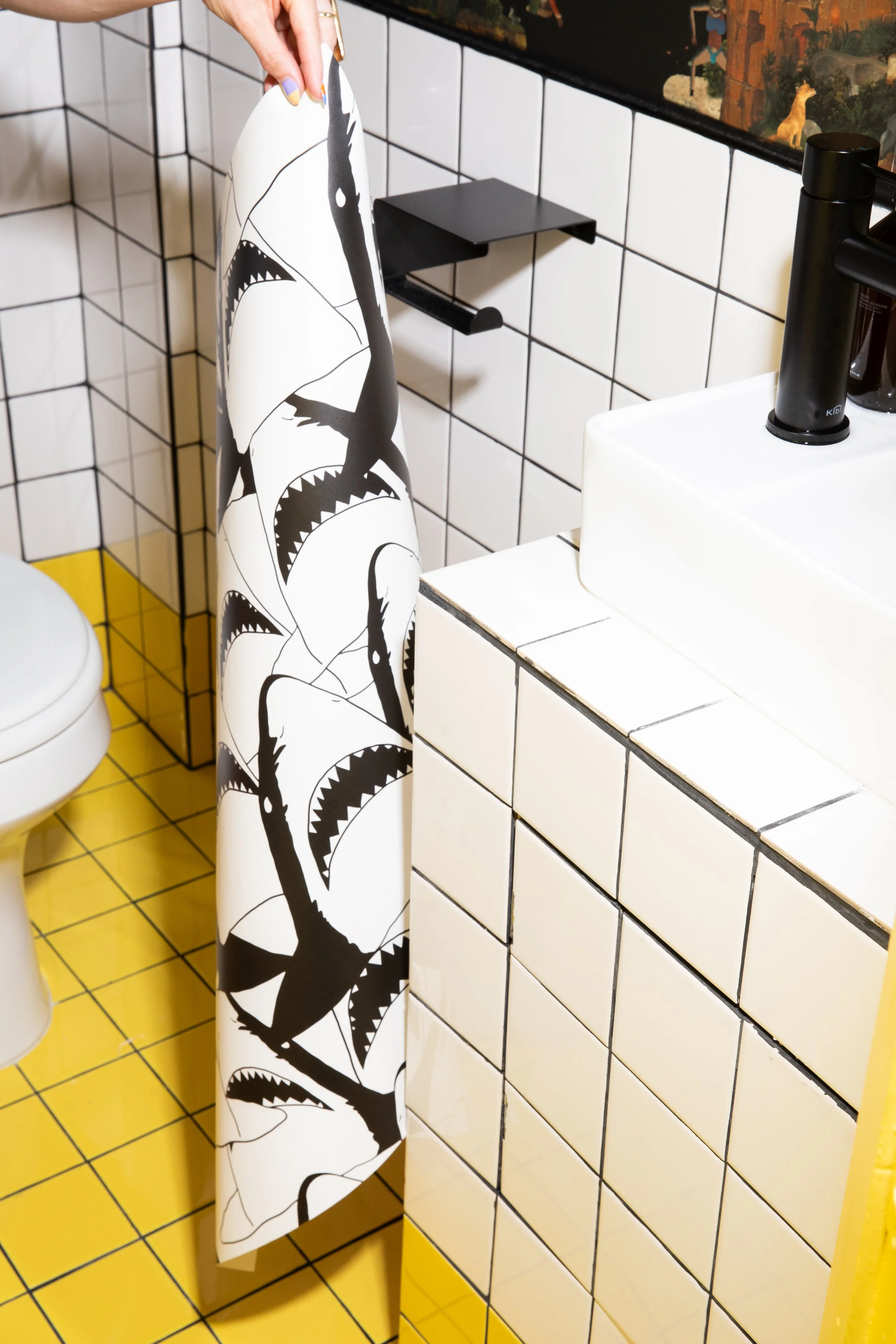 Black-and-white shark illustration wallpaper held beside a tiled studio washroom sink with black fixtures and yellow floor tiles.