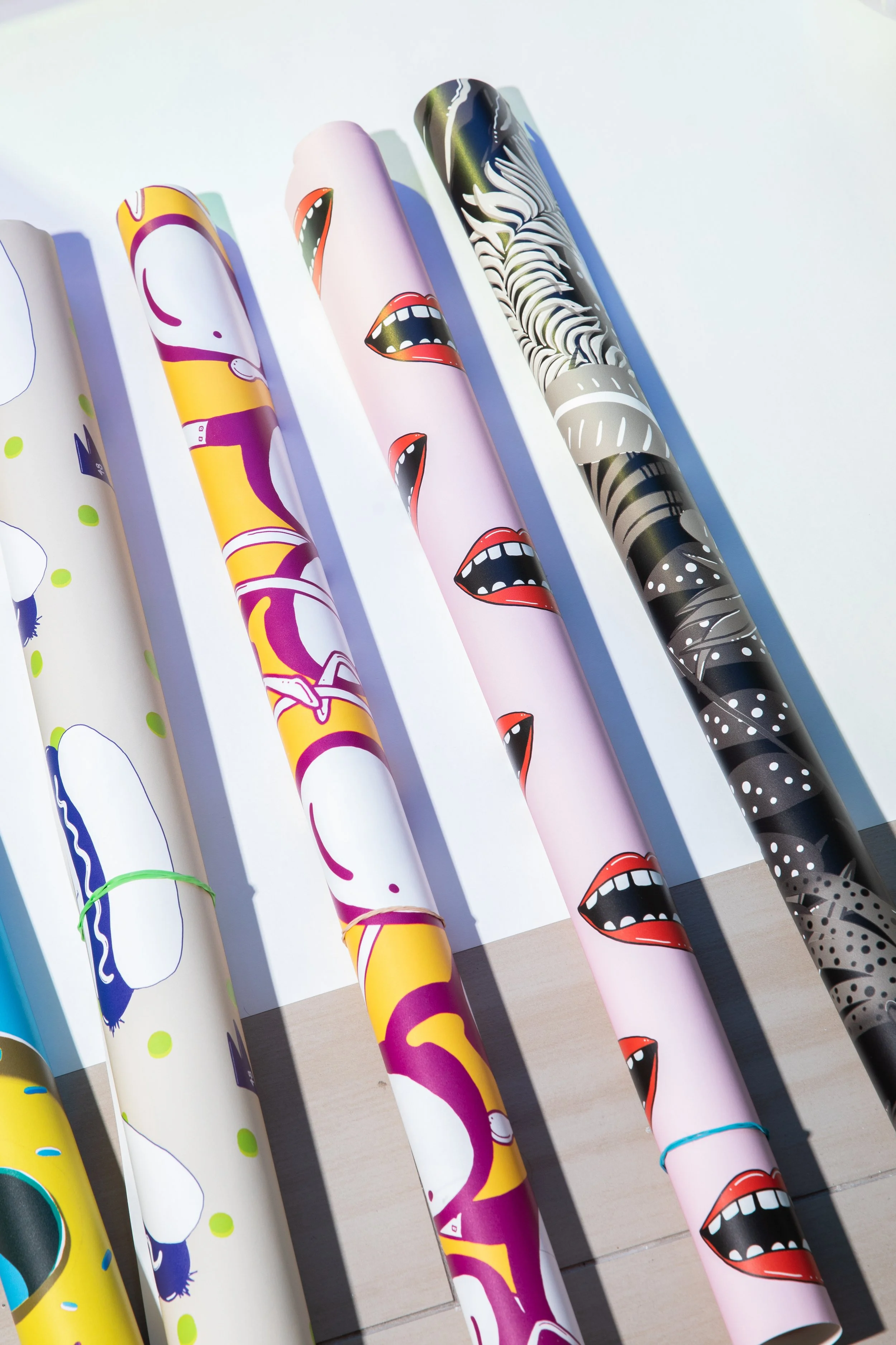 Colourful rolls of illustrated flodström wallpaper arranged diagonally in sunlight on a pale floor.