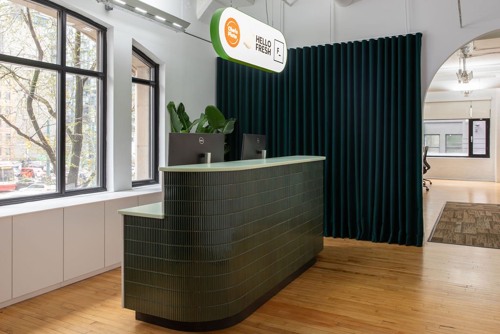 Toronto office reception designed by Flodström Interiors for HelloFresh, Chef's Plate and Factor, featuring a curved green tiled desk, dark green curtains and suspended brand signage.