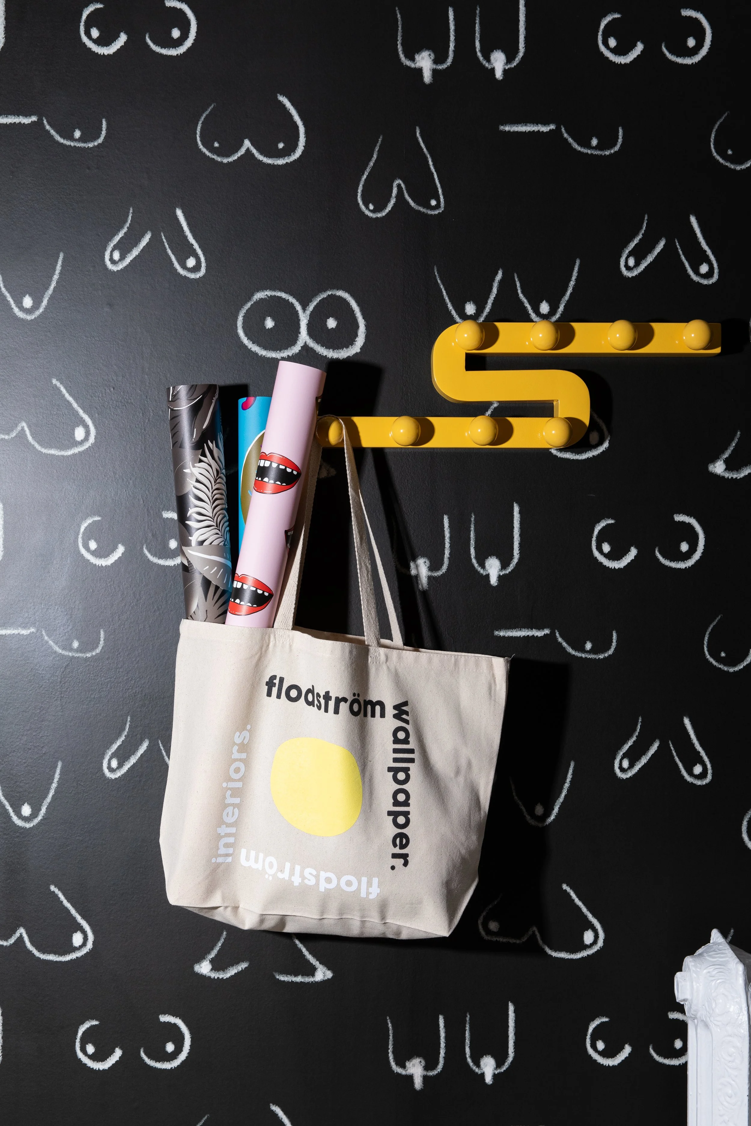 Flodström wallpaper rolls in a branded canvas tote hanging against a black wall with white illustrated motifs.