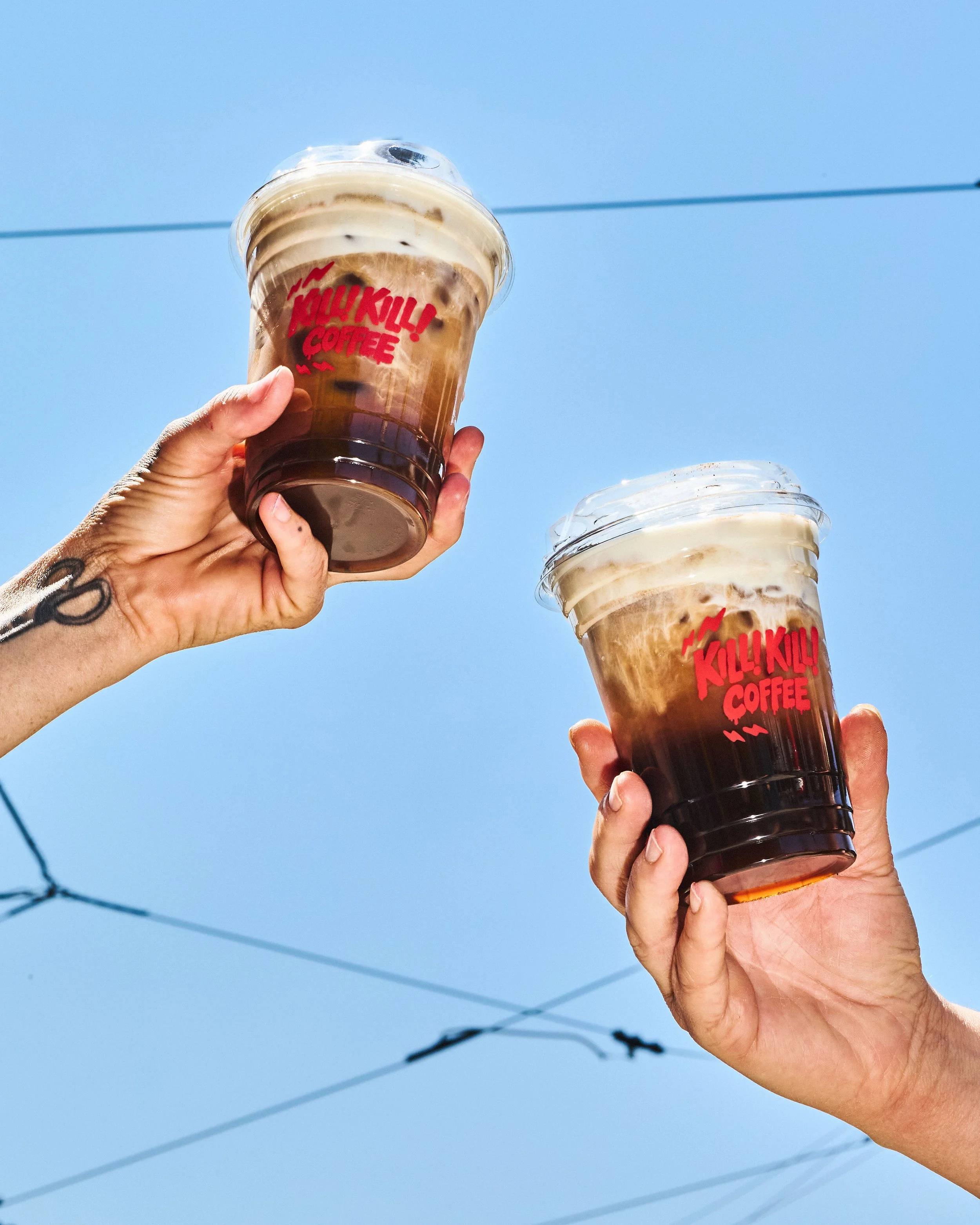 Two clear iced coffee cups featuring red Kill! Kill! Coffee logos, held against a bright blue sky.
