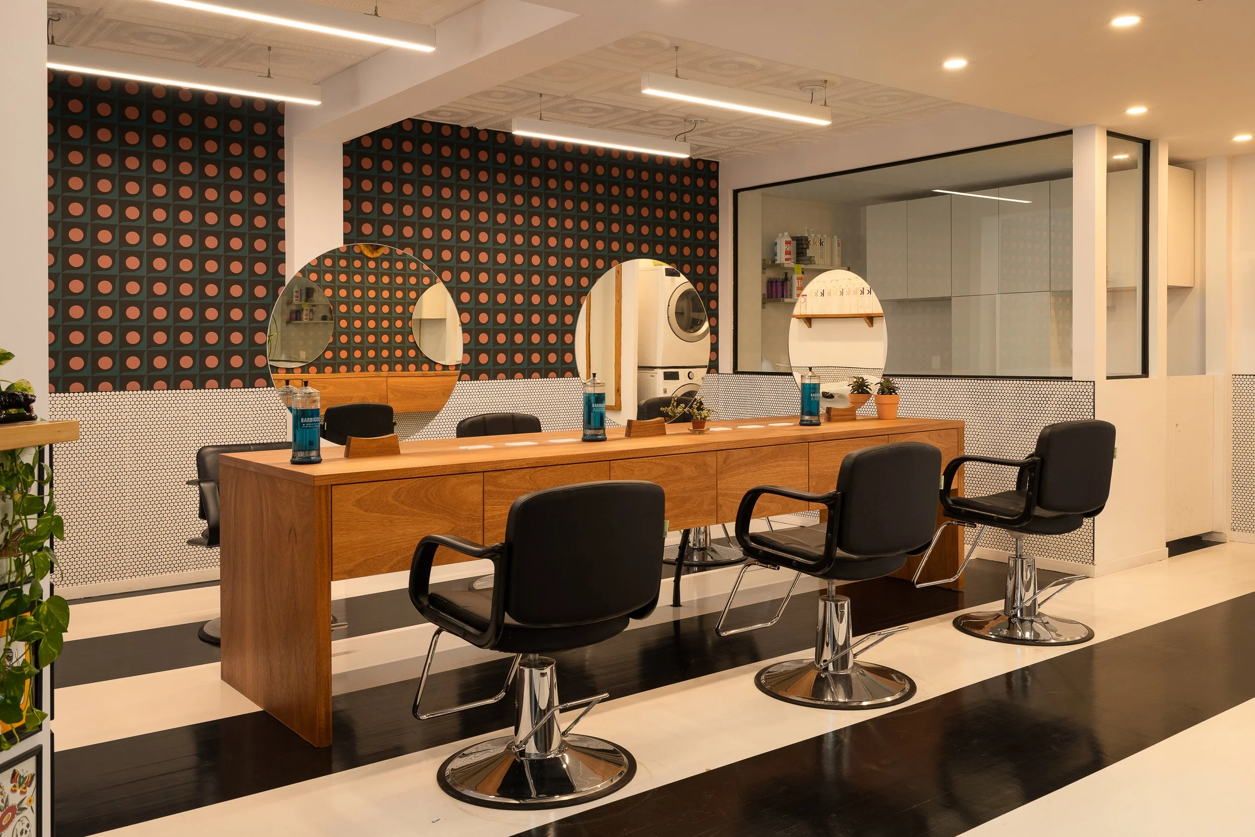 Interior of Two Horses hair salon in Montreal, featuring black-and-white striped floors, custom wood styling stations, circular mirrors and geometric wallpaper.