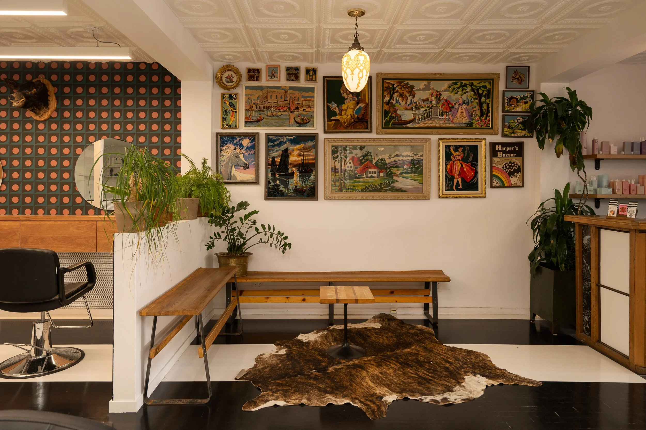 Salon waiting area with wooden benches, vintage needlepoint artwork, potted plants and a cowhide rug over painted striped floors.
