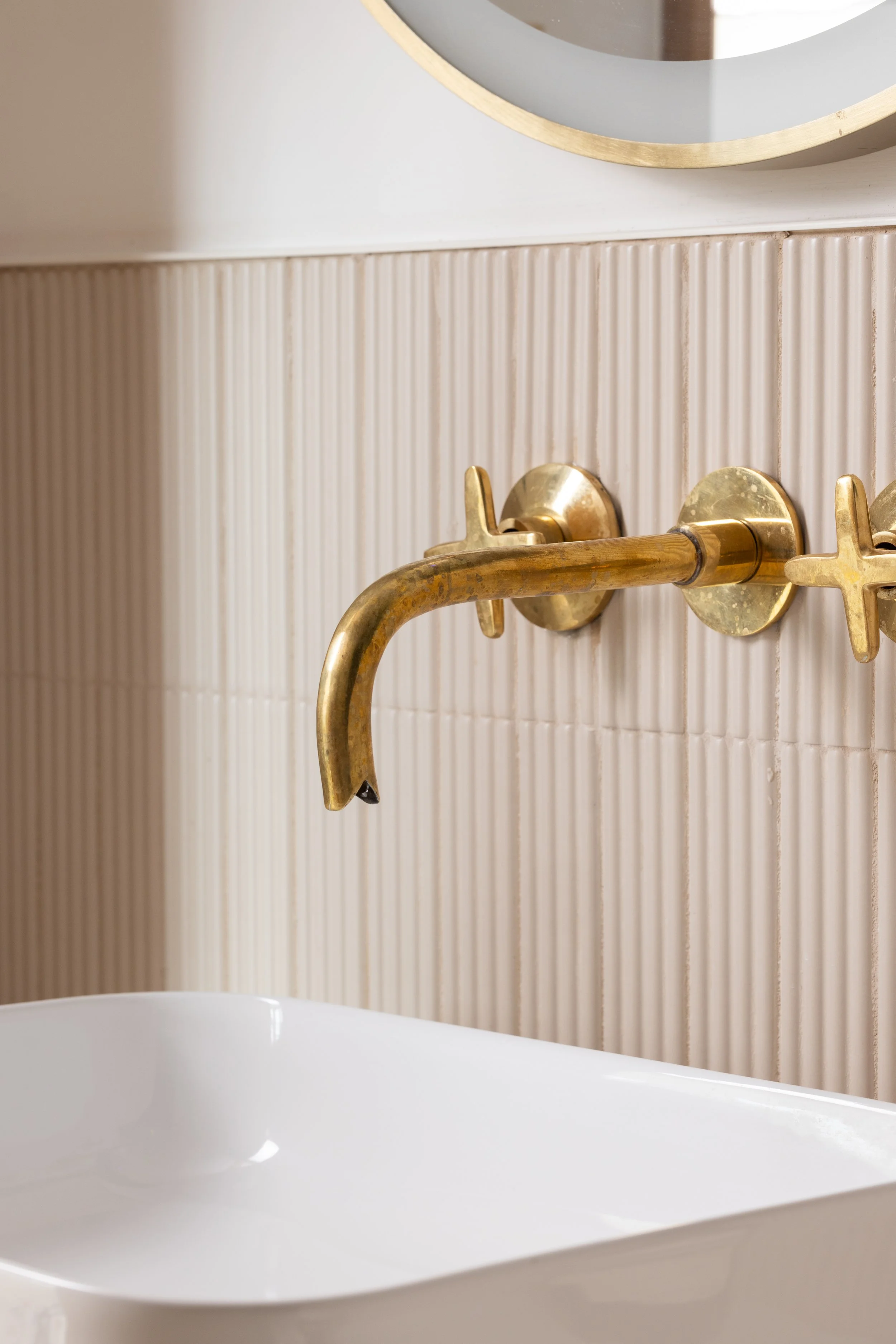 Close-up of an aged brass wall-mounted faucet against pale vertically fluted bathroom tile.