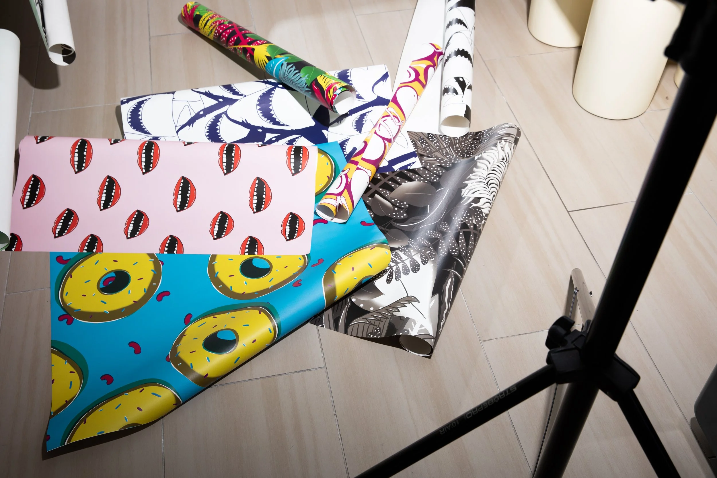 Assorted flodström wallpaper rolls and sheets arranged on a pale floor, featuring doughnuts, mouths, sharks and botanical illustrations.