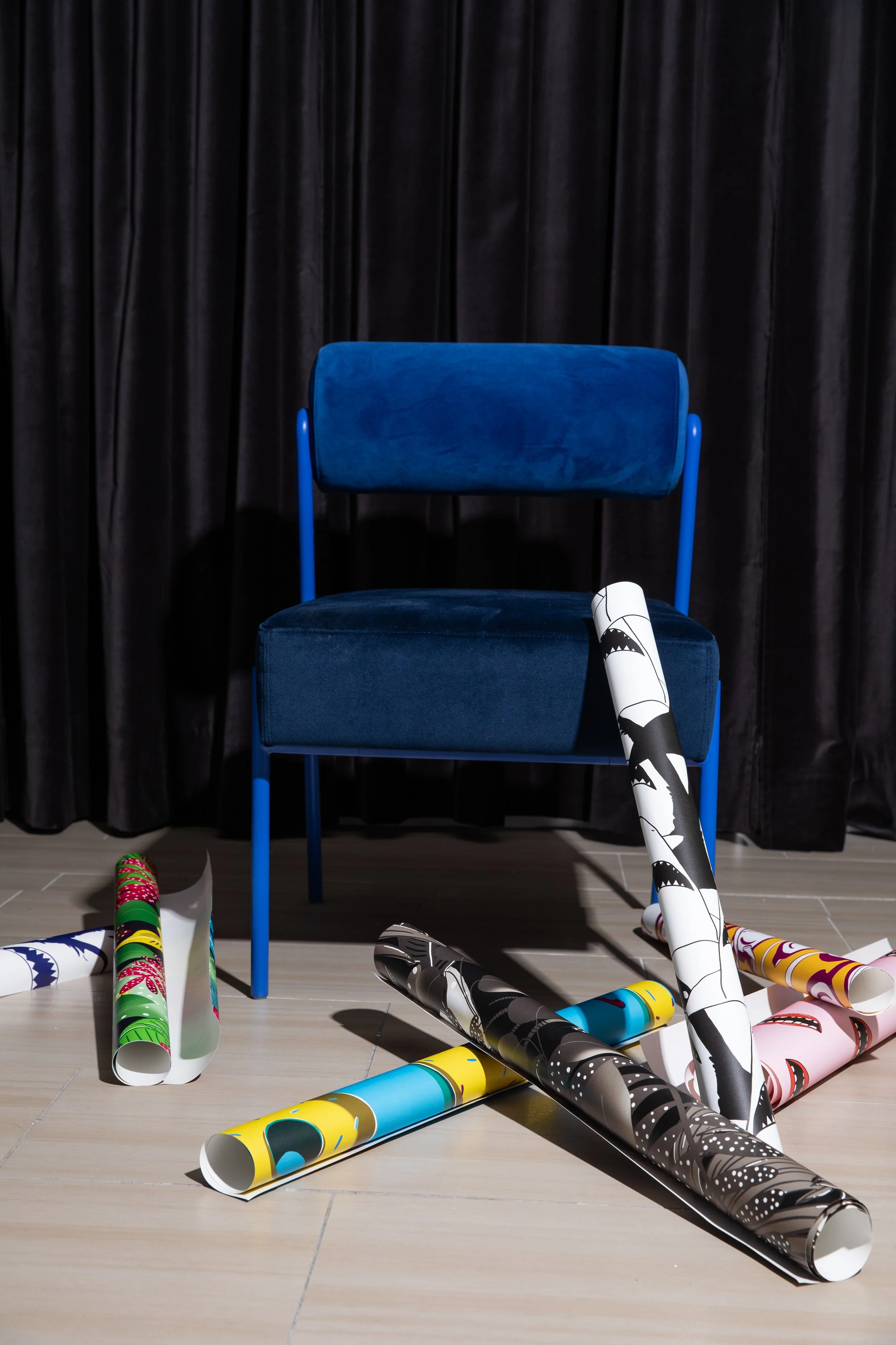 Cobalt blue upholstered chair against black curtains with colourful rolls of illustrated wallpaper scattered across the floor.