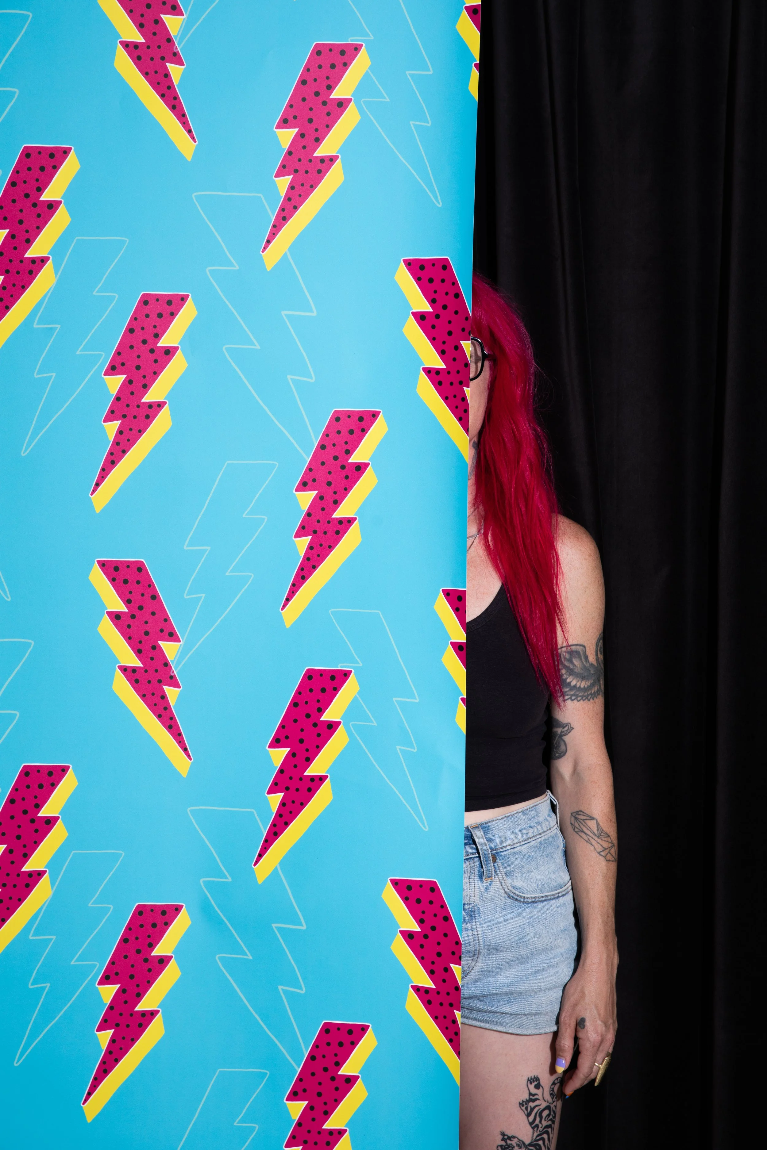 Designer partially concealed behind bright blue wallpaper featuring pink and yellow lightning bolts against a black curtain.