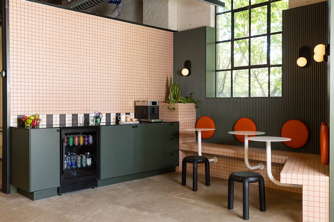 Office café and lounge designed by Flodström Interiors for Farmhouse Creative Labs in Toronto, featuring pale pink tiled walls, dark green cabinetry, orange upholstered seating and custom café tables.