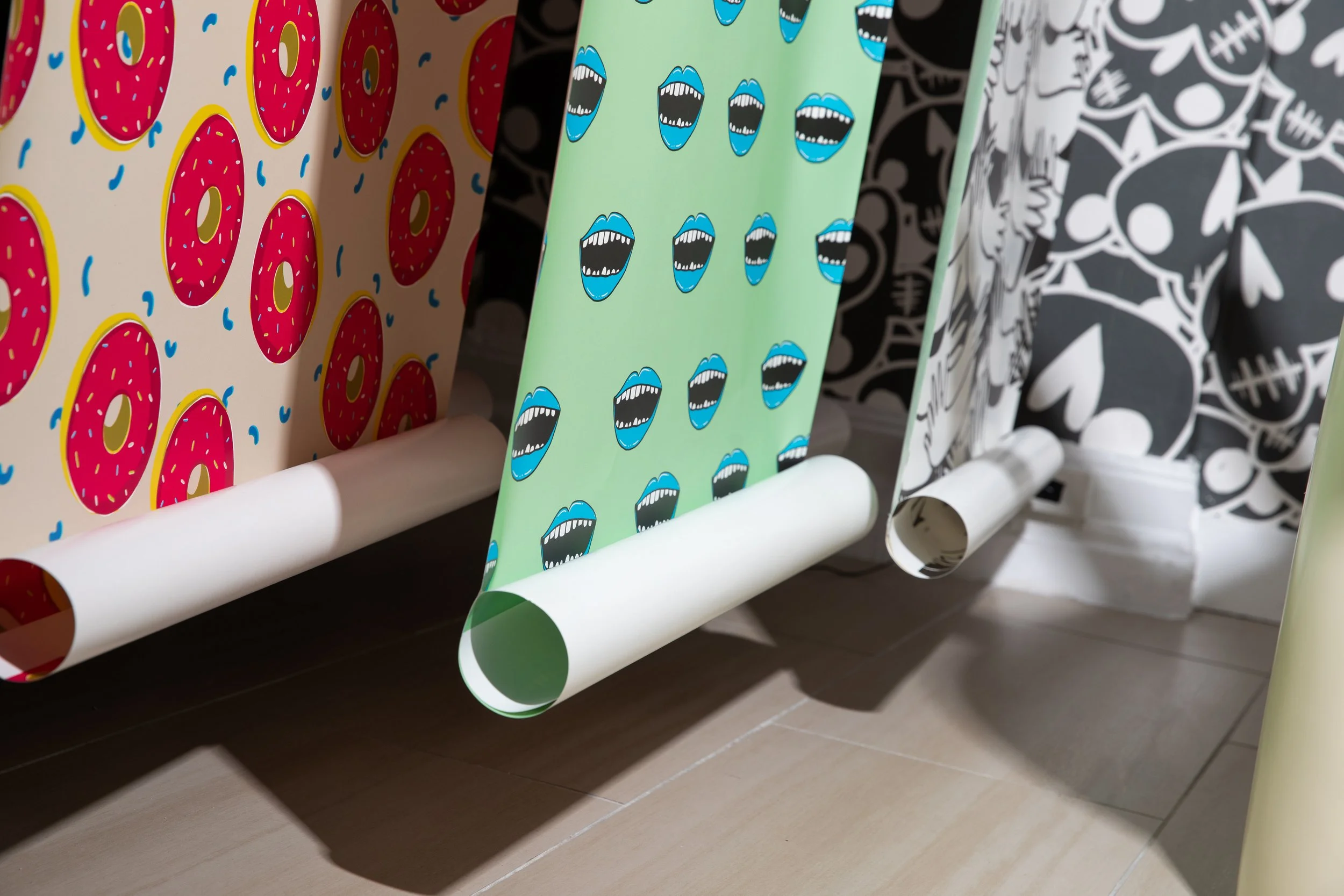 Hanging wallpaper rolls featuring pink doughnuts, blue mouths and black-and-white illustrated patterns.