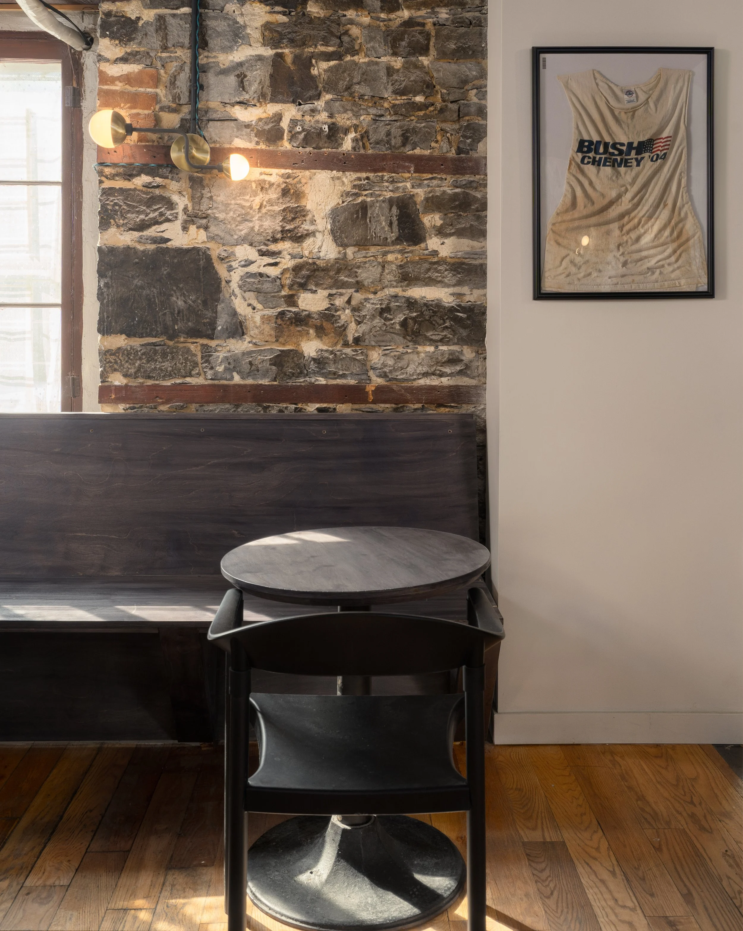 Small round table and dark wood banquette beside an exposed stone wall and framed memorabilia.